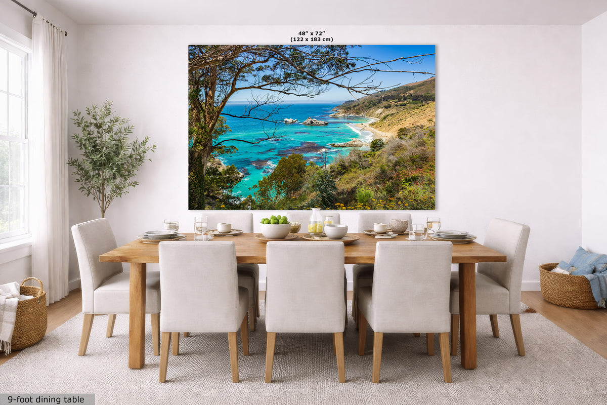 A large wall art print showing a coastal landscape with turquoise water, sandy beach, rocky shoreline, and trees, displayed above a dining table in a bright modern room. The print includes size text at the top and a small label at bottom left.