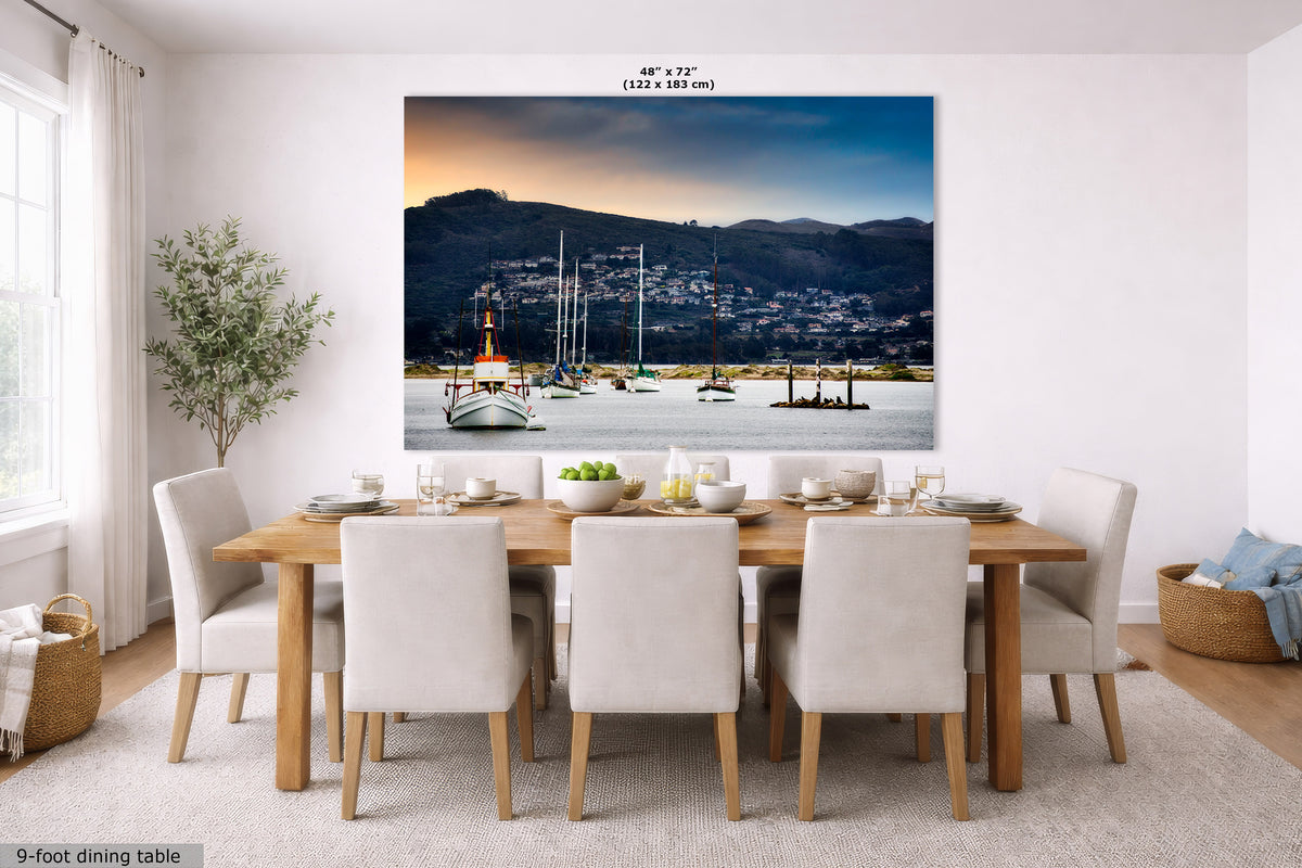 A large wall mural or canvas print depicting a coastal harbor scene with boats, mountains, and a sunset, displayed above a dining table in a bright modern room. The print has text at the top reading 48" x 72" and a small label at bottom left reading 9-foot dining table.