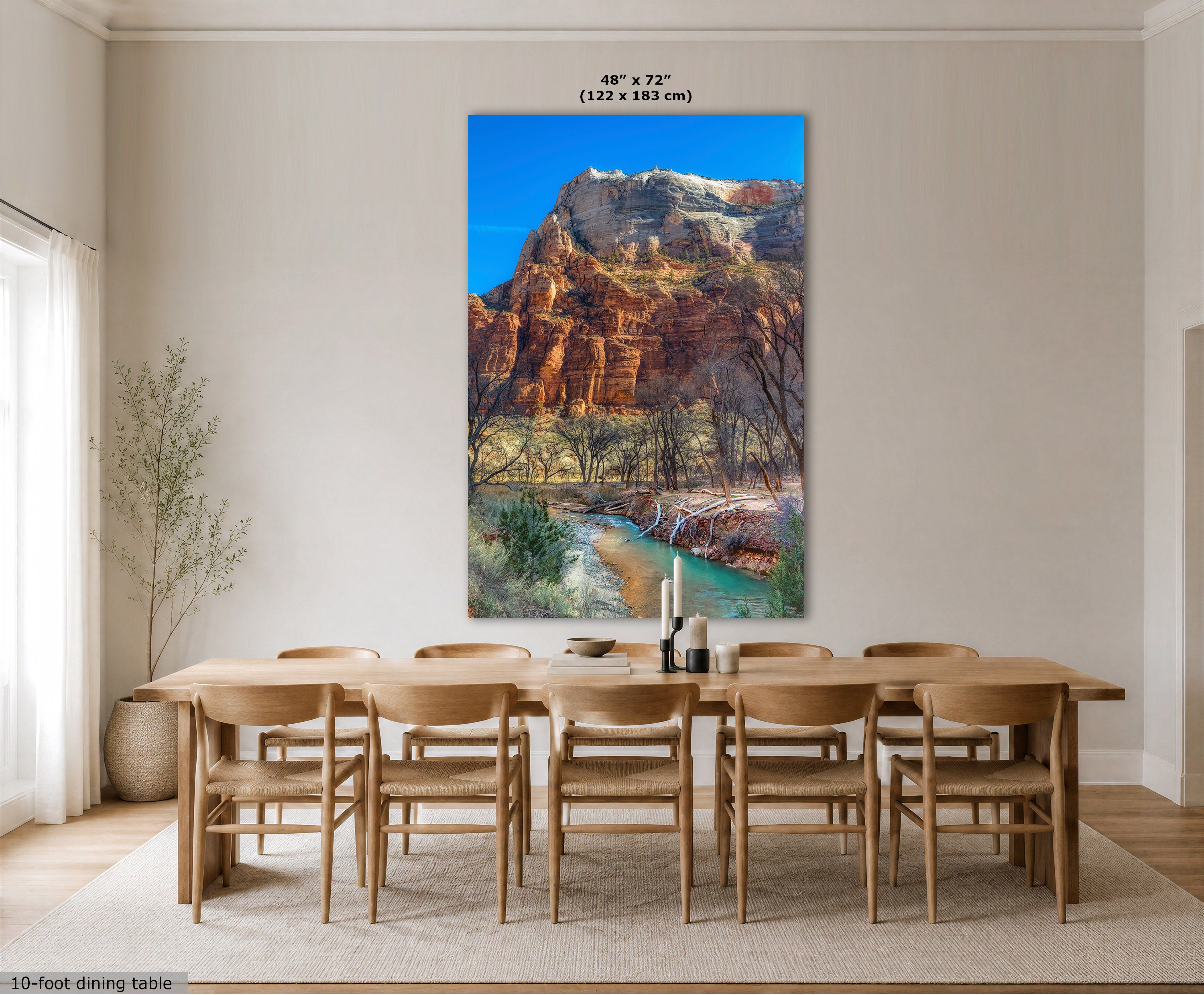 A large vertical wall art print showing a canyon landscape with red rock cliffs, a turquoise river, and blue sky, displayed above a wooden dining table and chairs in a bright neutral dining room. Text at the top reads 48 x 72" (122 x 183 cm), and a small note at bottom left says 10-foot dining table.