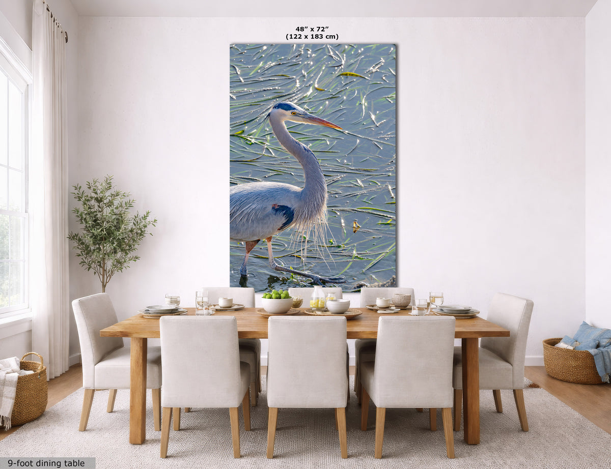 A large vertical wall art print showing a blue heron standing in shallow water with reeds, displayed above a dining table in a bright, modern dining room. The print includes size text at the top and a small caption at the bottom left.