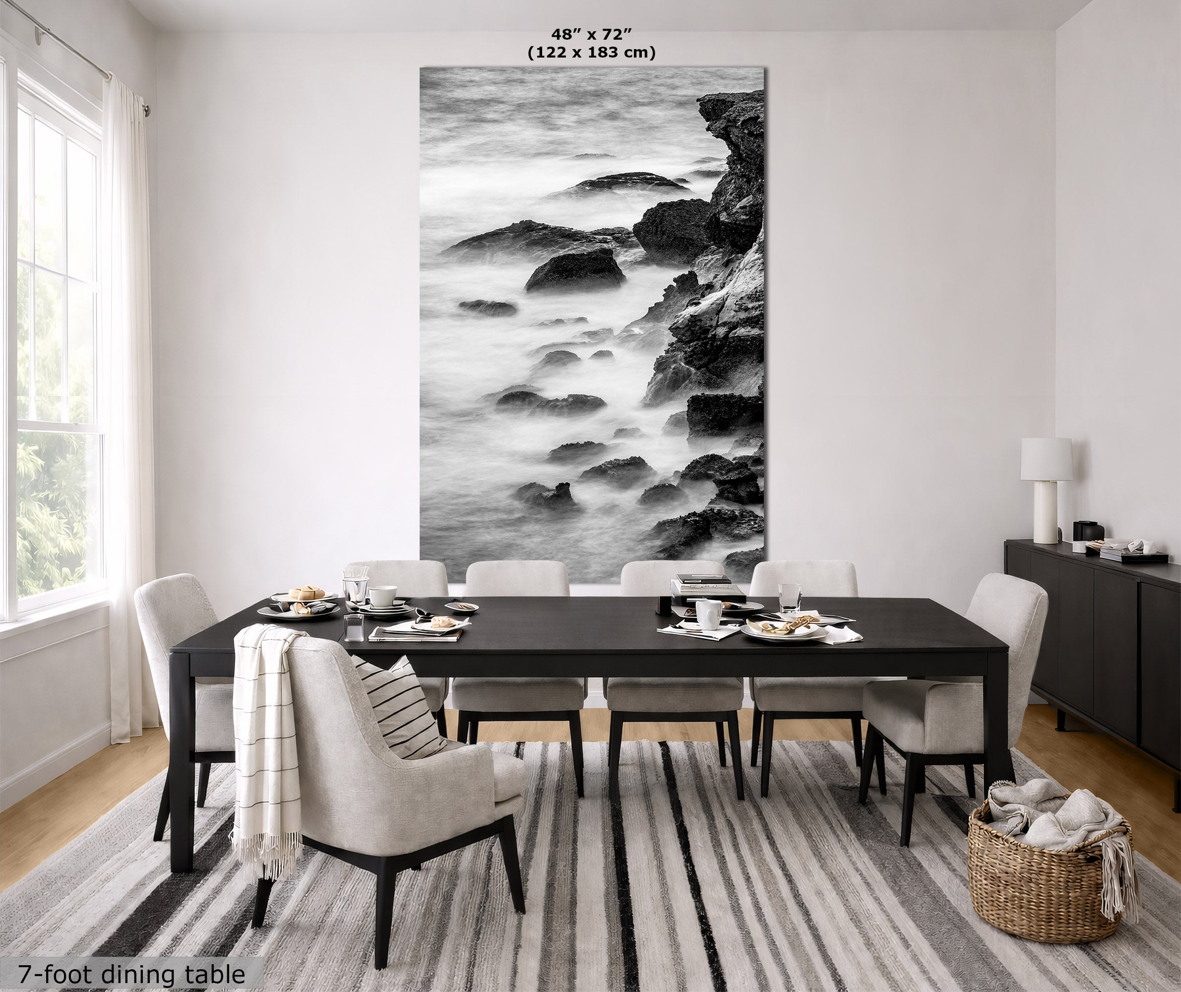 A large black and white vertical wall art print depicting a rocky shoreline with long-exposure water, displayed above a dark dining table in a bright modern dining room. Text at the top reads 48" x 72" (122 x 183 cm) and text at the bottom left reads 7-foot dining table.