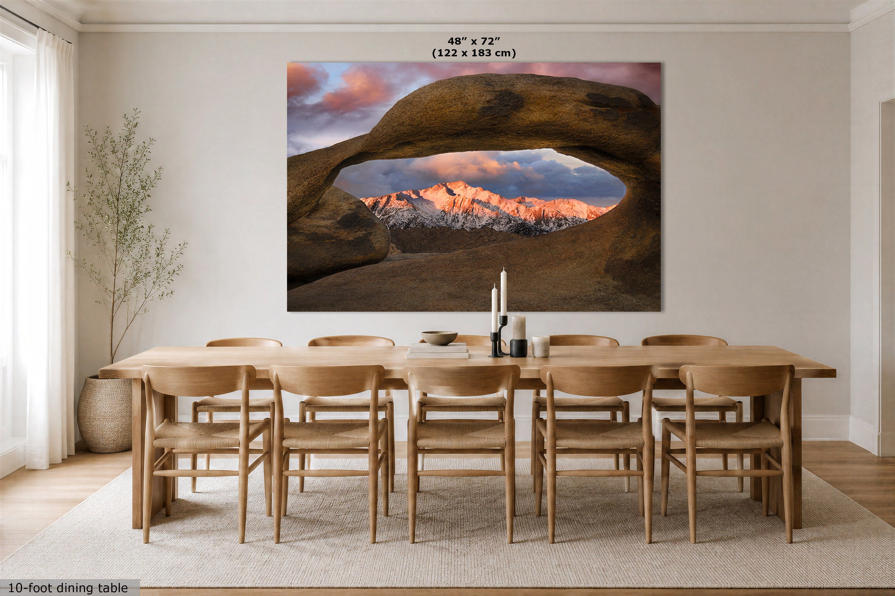A large wall art print showing a desert rock arch framing snow-capped mountains at sunset, displayed above a wooden dining table and chairs in a bright neutral dining room. Text at the top reads 48" x 72" (122 x 183 cm), and a small note at bottom left says 10-foot dining table.