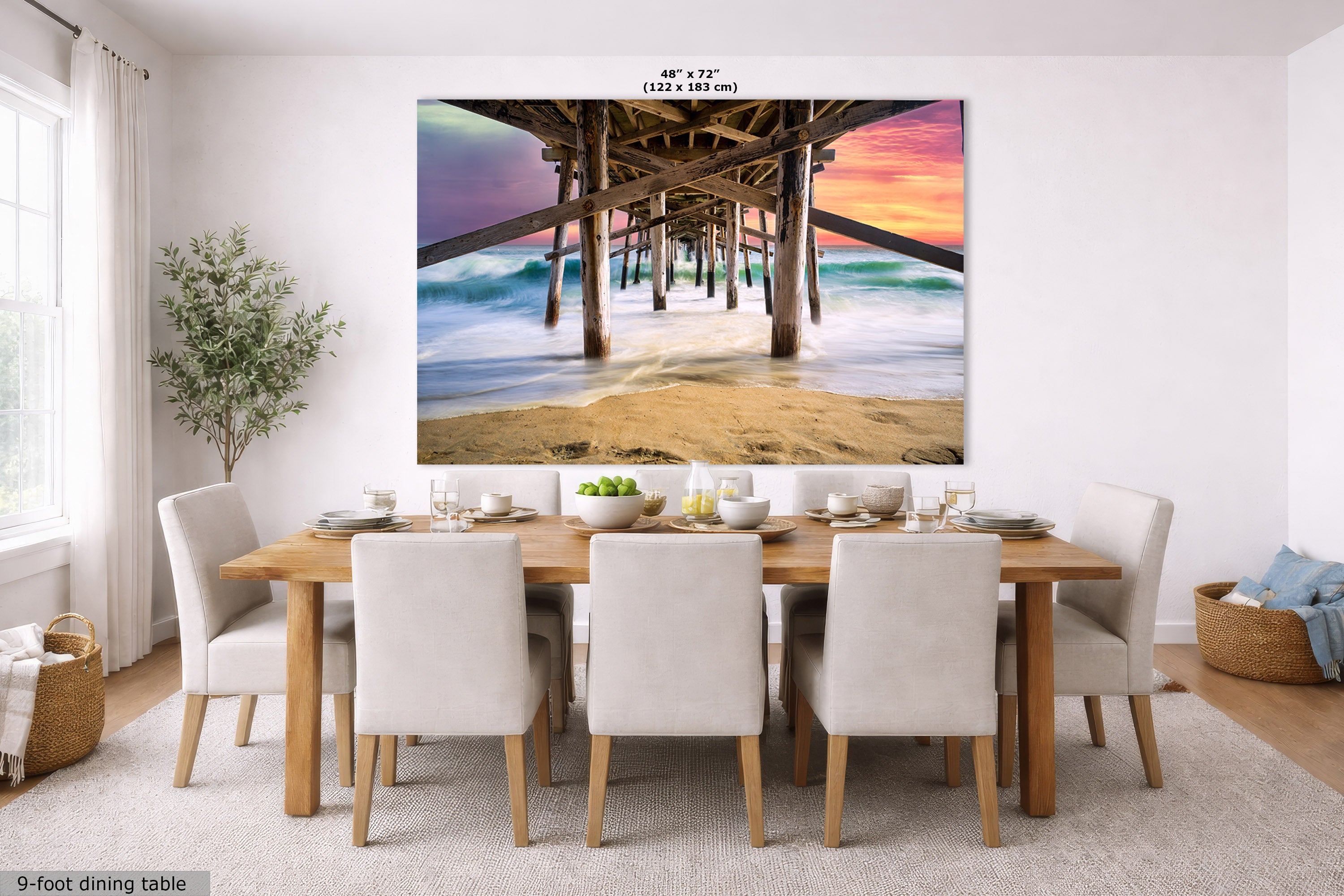 A large rectangular wall art print showing a pier overlooking a beach at sunset, displayed above a dining table in a bright modern room. The print includes size text at the top and a small label at bottom left reading 9-foot dining table.