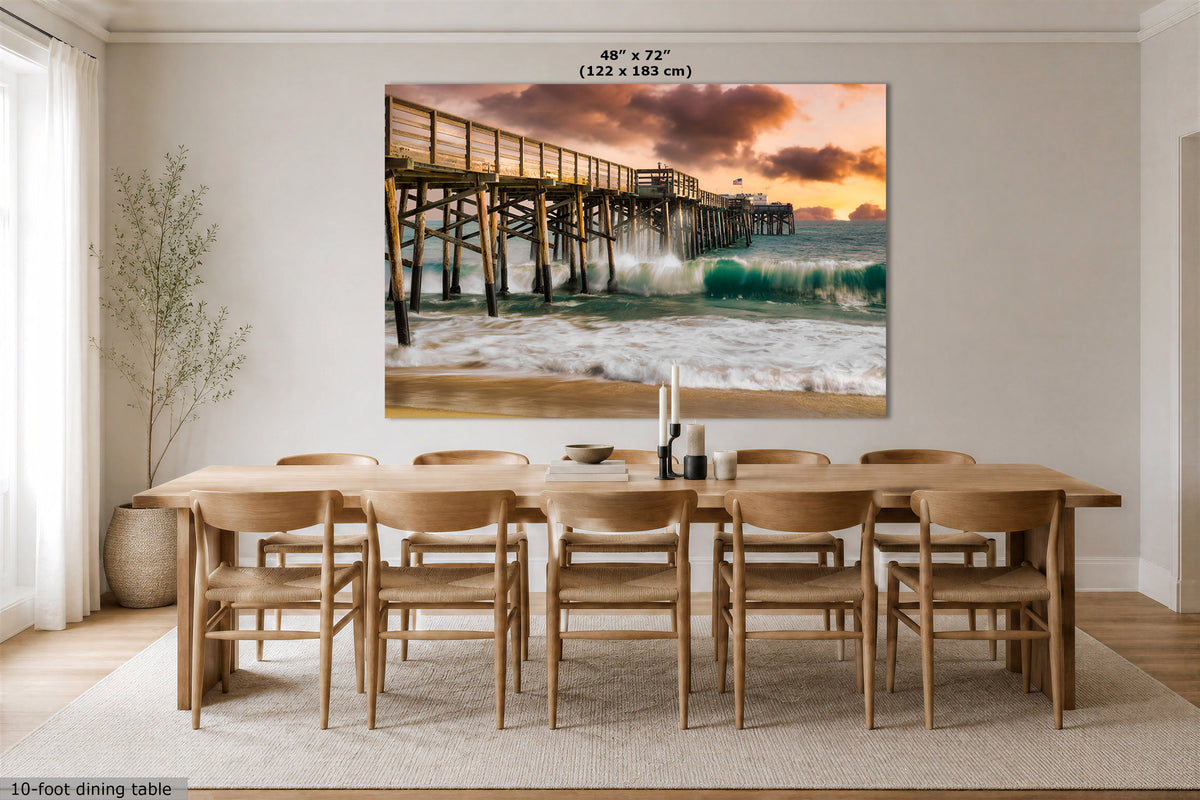 A large rectangular wall art print showing a pier over ocean waves at sunset, displayed above a wooden dining table and chairs in a bright dining room. The print includes size text at the top and a small note at bottom left about the 10-foot dining table.