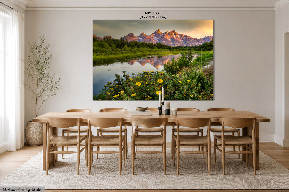 A large rectangular wall art print showing a mountain landscape with a reflective river and wildflowers, displayed above a wooden dining table and chairs in a bright dining room. Small text at the top reads 48" x 72" (122 x 183 cm), and a small caption at the bottom left says 10-foot dining table.