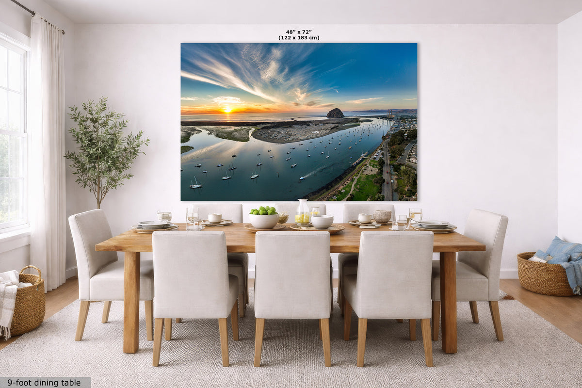 A large wall art print showing a coastal harbor sunset with boats and clouds, displayed above a dining table in a bright modern room. The print includes size text at the top and a small label at bottom left reading 9-foot dining table.