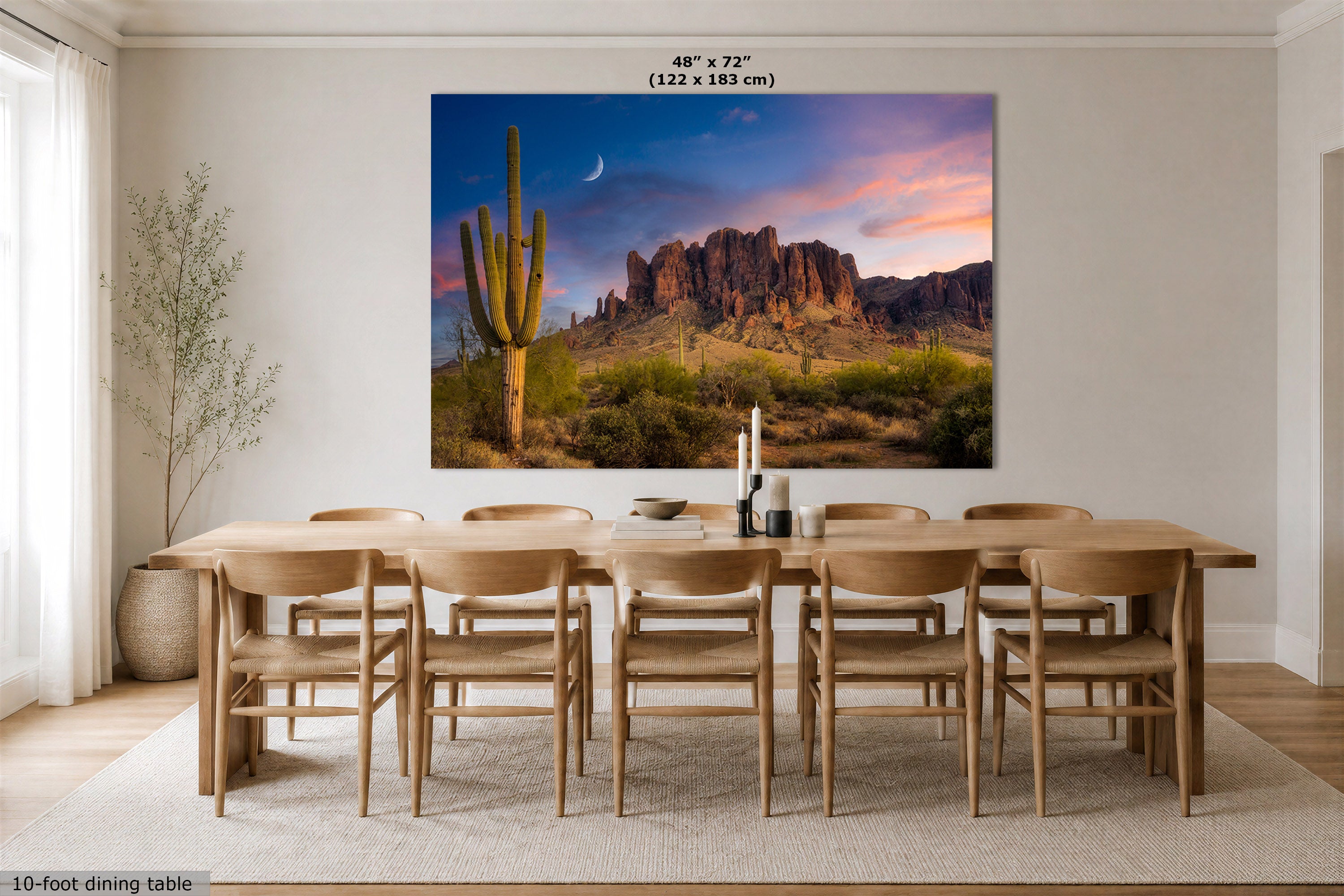 A large wall art print showing a desert landscape with a saguaro cactus, rocky mountains, and a pink-purple sky, displayed above a wooden dining table and chairs in a bright dining room. Text at the top reads 48" x 72" (122 x 183 cm).
