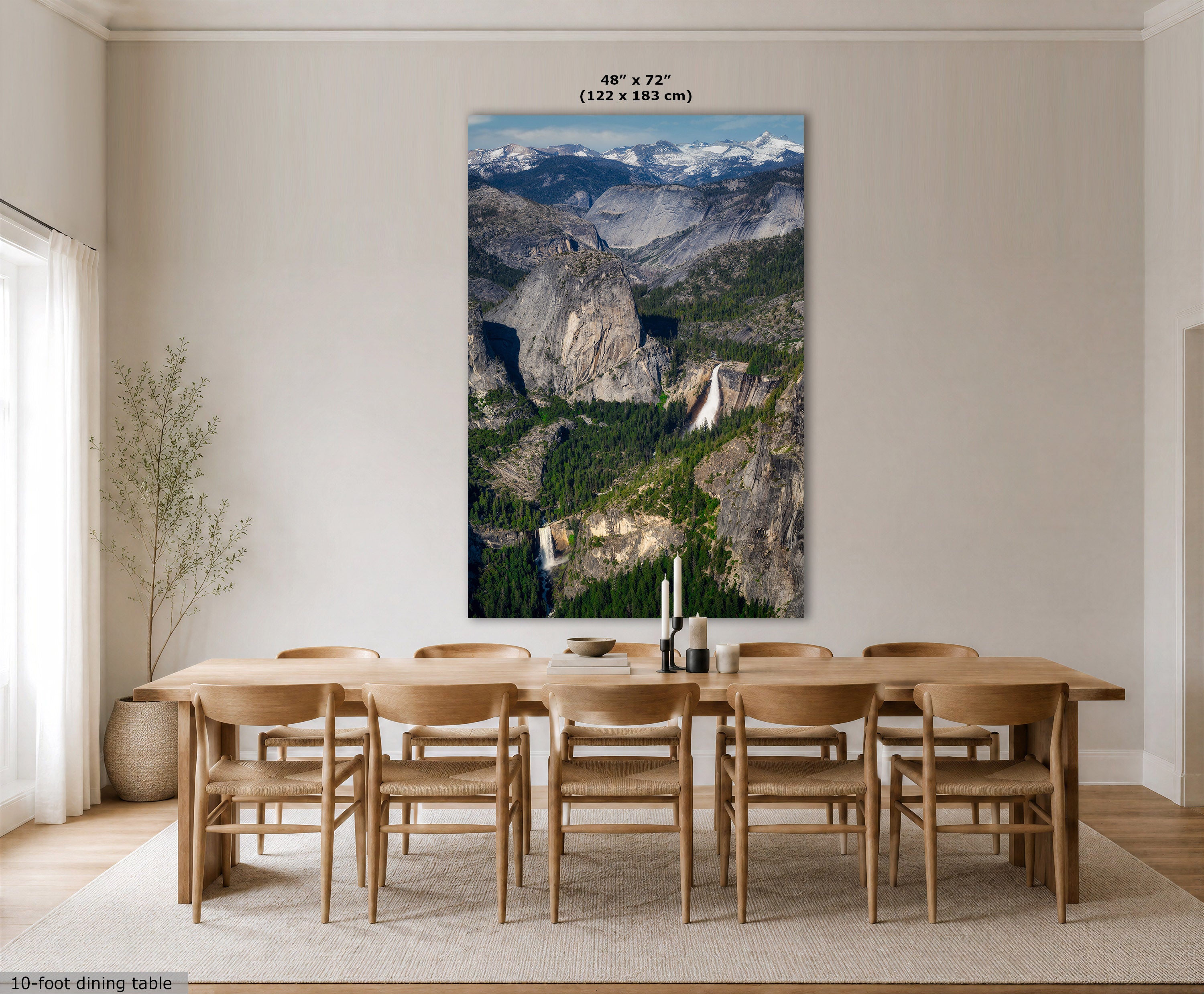 A large vertical wall art print showing a mountain valley landscape with waterfalls, displayed above a wooden dining table and chairs in a bright neutral dining room. The print includes size text at the top and a small note at bottom left about the 10-foot dining table.