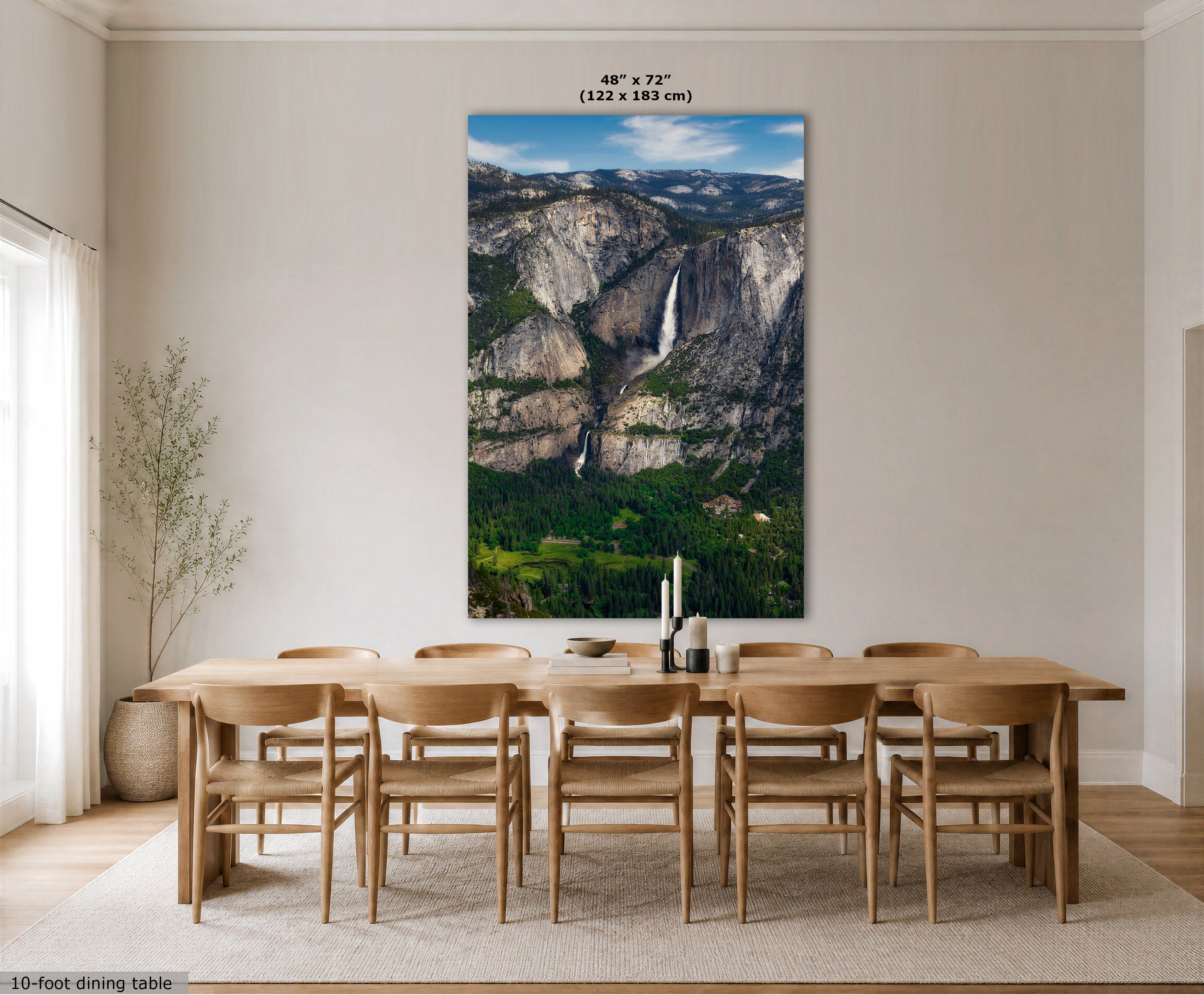 A large vertical Yosemite Falls mountain art print showing a mountain landscape with a waterfall, displayed above a wooden dining table and chairs in a bright dining room. The print includes size text at the top and a small label at the bottom left.
