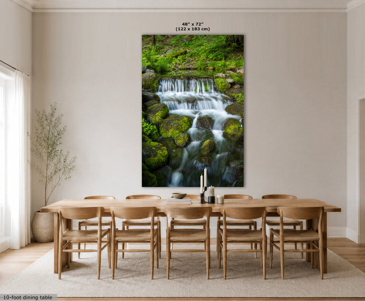 A large vertical mossy waterfall wall art print showing a waterfall over mossy rocks, displayed above a wooden dining table and chairs in a bright, modern dining room. The print includes size text at the top and a small label at the bottom left.