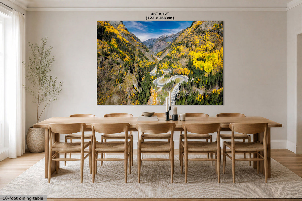 A large wall art print showing a mountain valley with yellow trees and a winding road, displayed above a wooden dining table and chairs in a bright dining room. Text at the top reads 48" x 72" (122 x 183 cm), and text at the bottom left says 10-foot dining table.