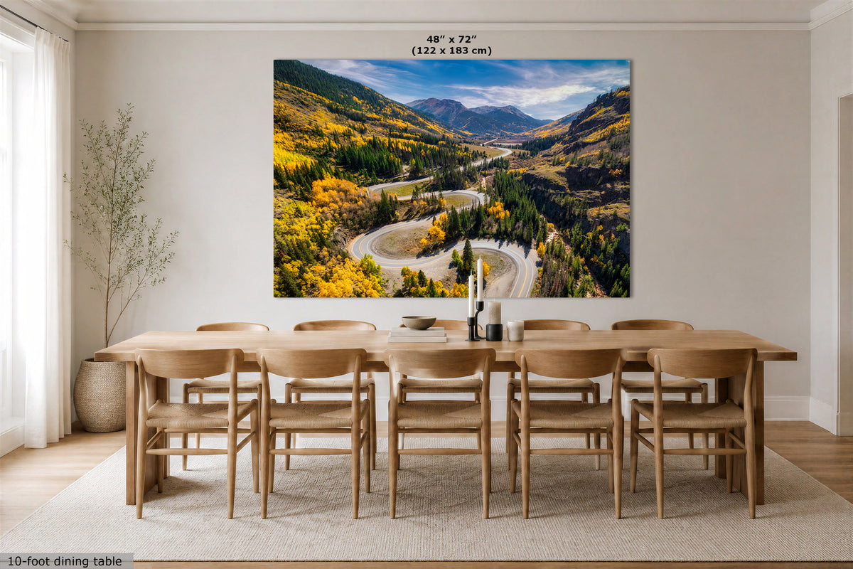 A large wall art print showing a winding mountain road through autumn-colored forests hangs above a wooden dining table and chairs in a bright dining room. The print includes a small size label reading 48 in x 72 in (122 x 183 cm).