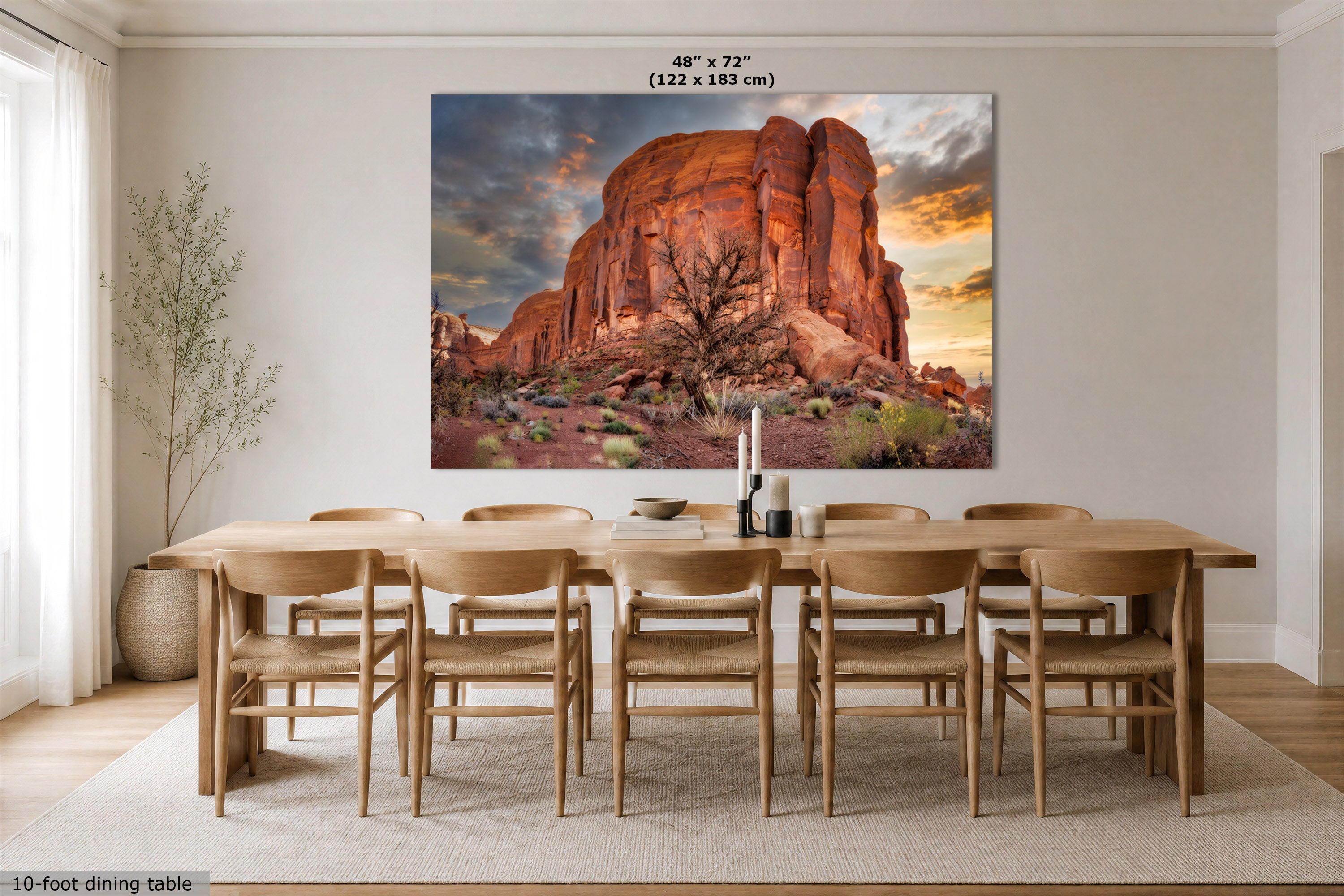 A large wall art print showing a desert rock formation at sunset, displayed above a wooden dining table and chairs in a bright dining room. Text at the top reads 48 x 72" (122 x 183 cm), and a small note at the bottom left says 10-foot dining table.