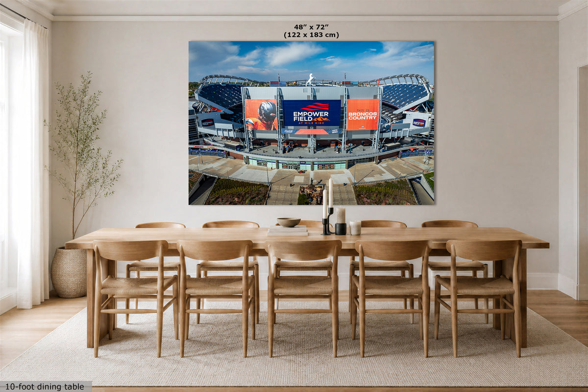 A Broncos Empower Field wall art print showing an aerial view of a football stadium under a blue sky, displayed above a wooden dining table in a bright modern room. Text at the top of the print reads 48" x 72" (122 x 183 cm), and the bottom-left corner shows a 10-foot dining table scale.