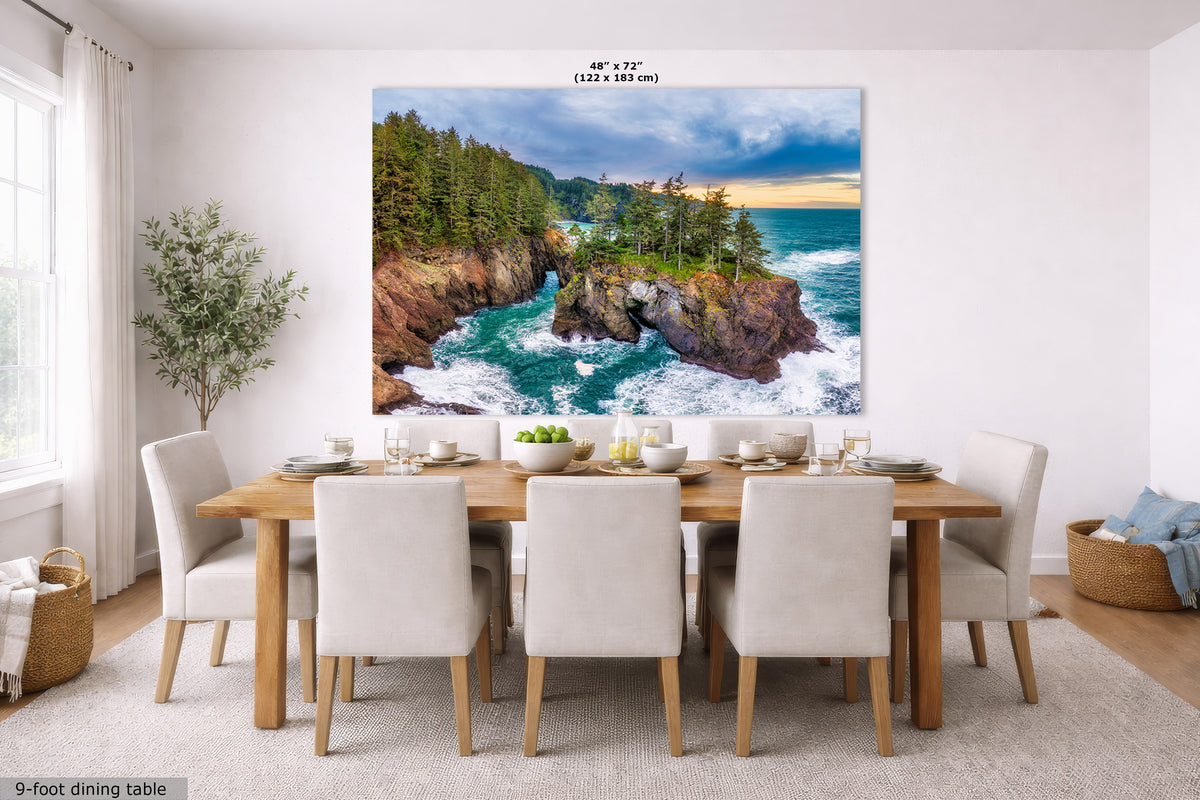 A large wall art print showing a coastal rocky shoreline with blue ocean water, green trees, and a cloudy sky, displayed above a dining table in a bright modern room. Text at the top reads 48" x 72" (122 x 183 cm), and text at the bottom left says 9-foot dining table.