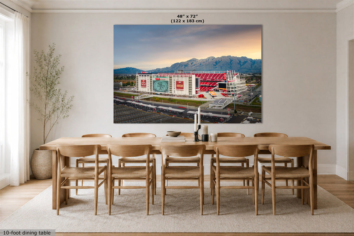 A large wall art print showing an outdoor stadium scene with mountains and a sunset sky, displayed above a wooden dining table and chairs in a bright modern room. Small text at the top reads 48" x 72" (122 x 183 cm), and text at the bottom left says 10-foot dining table.