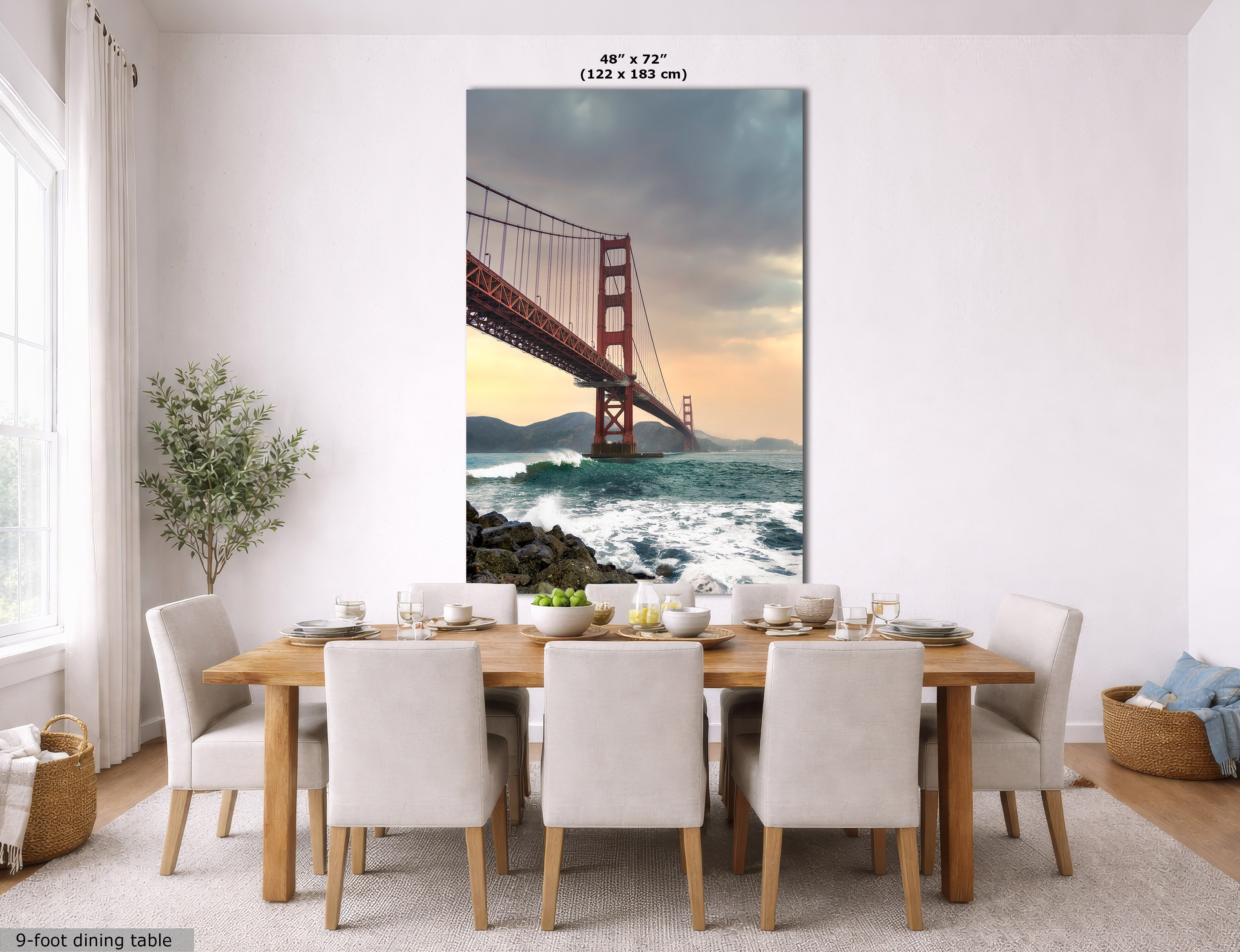 A large vertical wall art print of the Golden Gate Bridge over ocean waves is displayed above a dining table in a bright, modern dining room. The print shows the iconic red-orange bridge against a dramatic sky with clouds and warm sunset light.