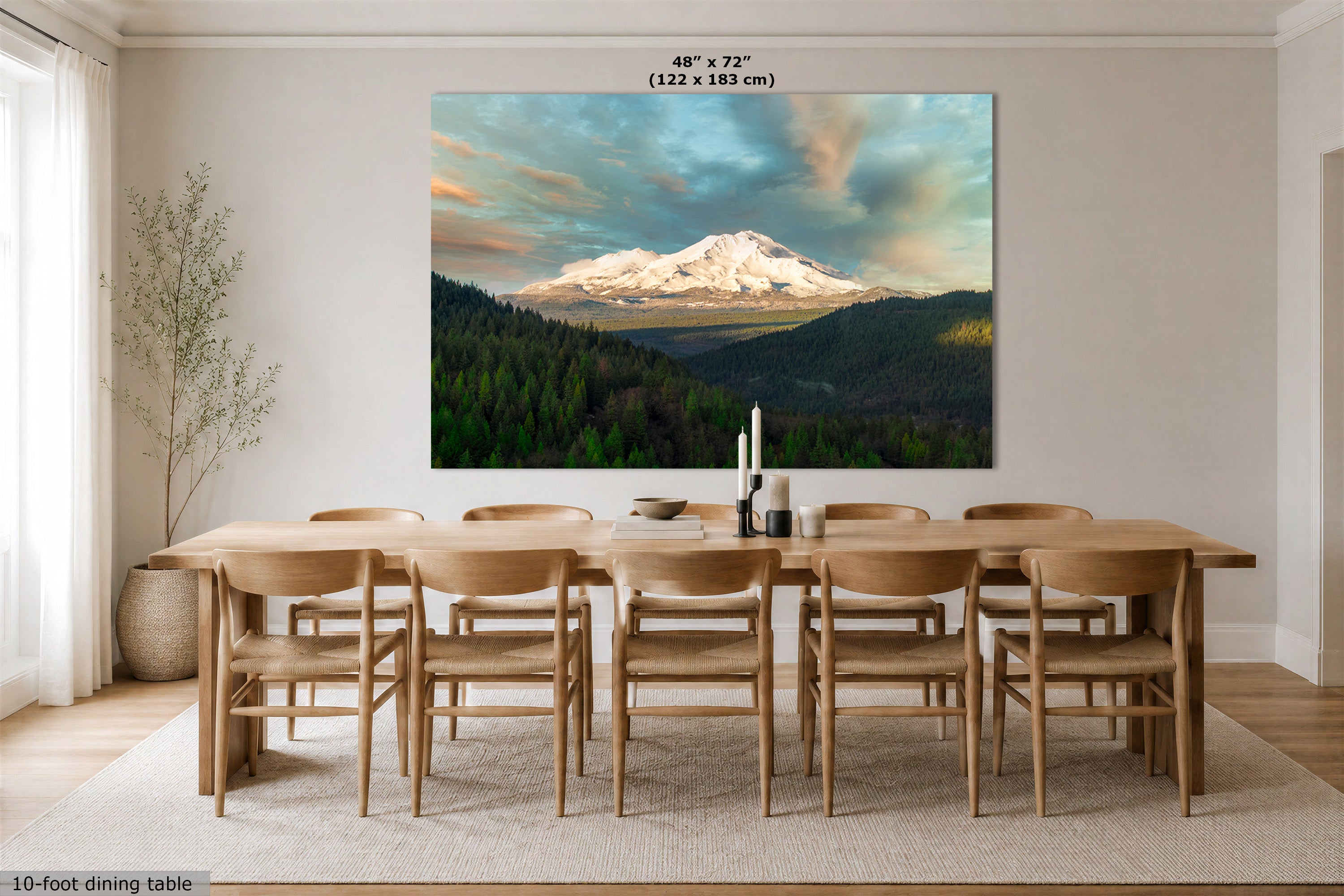 A large rectangular wall art print showing a snow-capped mountain landscape hangs above a wooden dining table and chairs in a bright dining room. The print includes size text at the top and a small note at the bottom left.