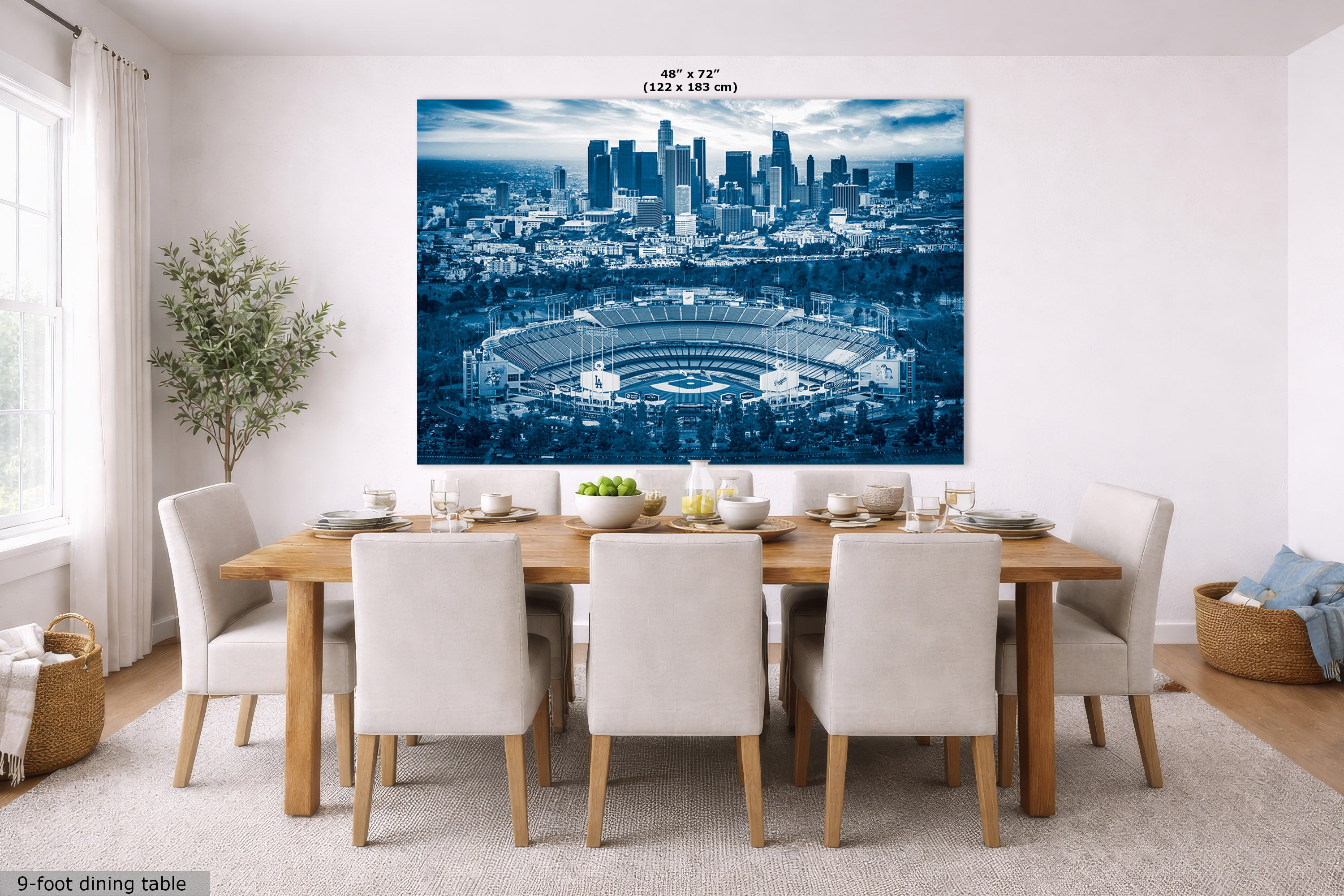 A large rectangular wall art print showing a blue-toned city skyline and stadium scene, displayed above a dining table in a bright modern room. Small text at the top reads 48" x 72" (122 x 183 cm), and text at bottom left says 9-foot dining table.