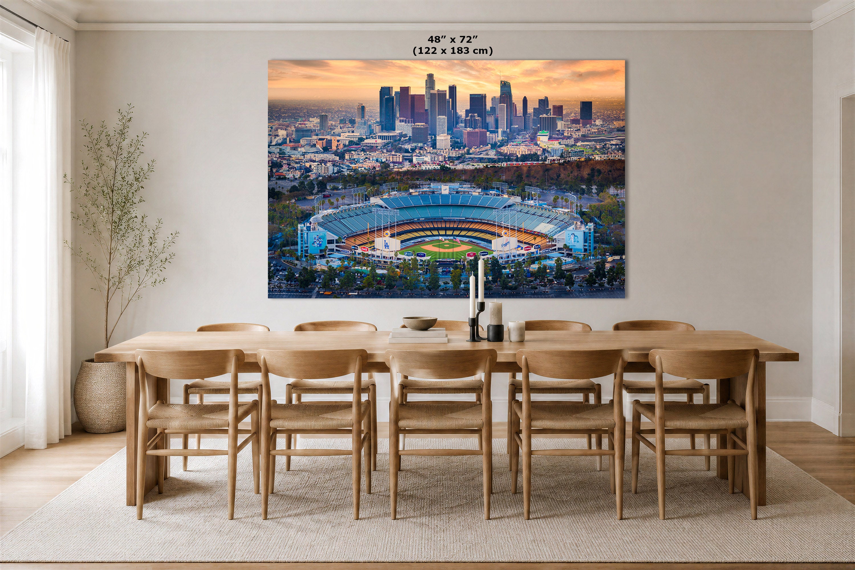 A large wall art print depicting an aerial view of a stadium and the Los Angeles skyline at sunset, displayed above a wooden dining table in a modern dining room. The print includes size text at the top reading 48" x 72" (122 x 183 cm).