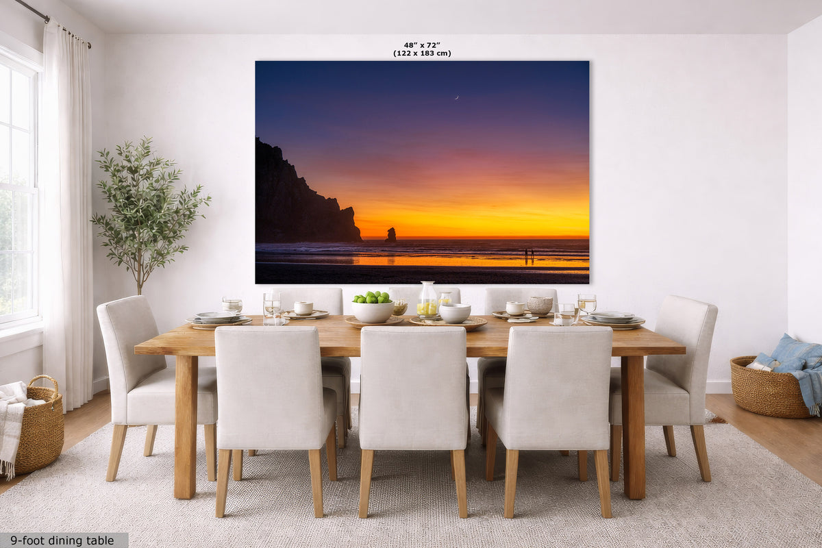 A large wall art print showing a coastal sunset scene hangs above a 9-foot dining table with white upholstered chairs in a bright, modern dining room. The print depicts a dramatic orange and purple sunset over the ocean with dark cliff silhouettes.