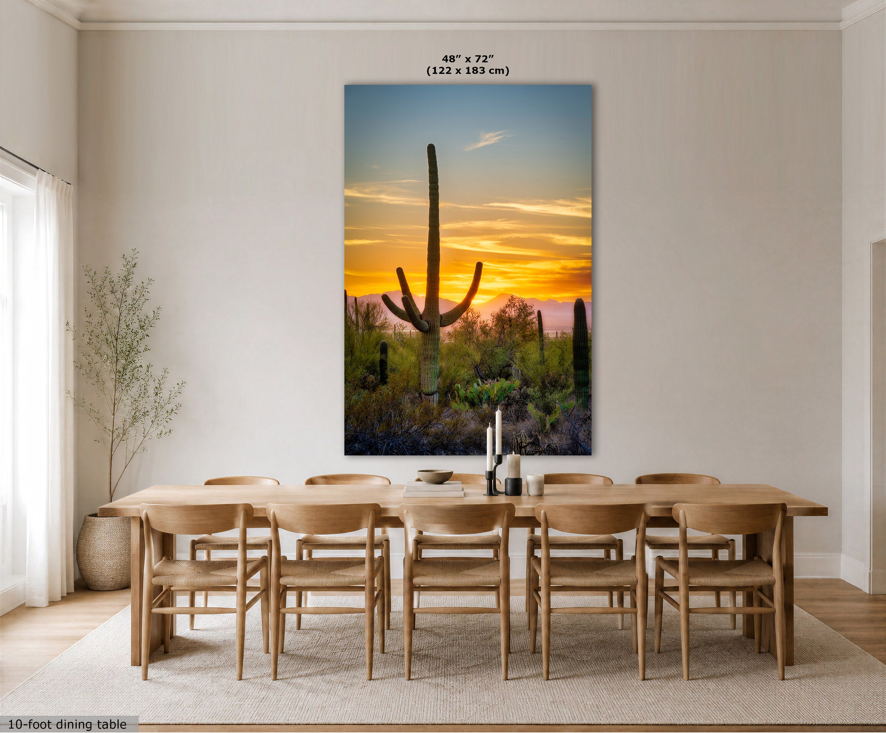 A large vertical wall art print showing a desert sunset with a saguaro cactus, displayed above a wooden dining table and chairs in a bright modern dining room. The print includes size text at the top and a small label at the bottom left.