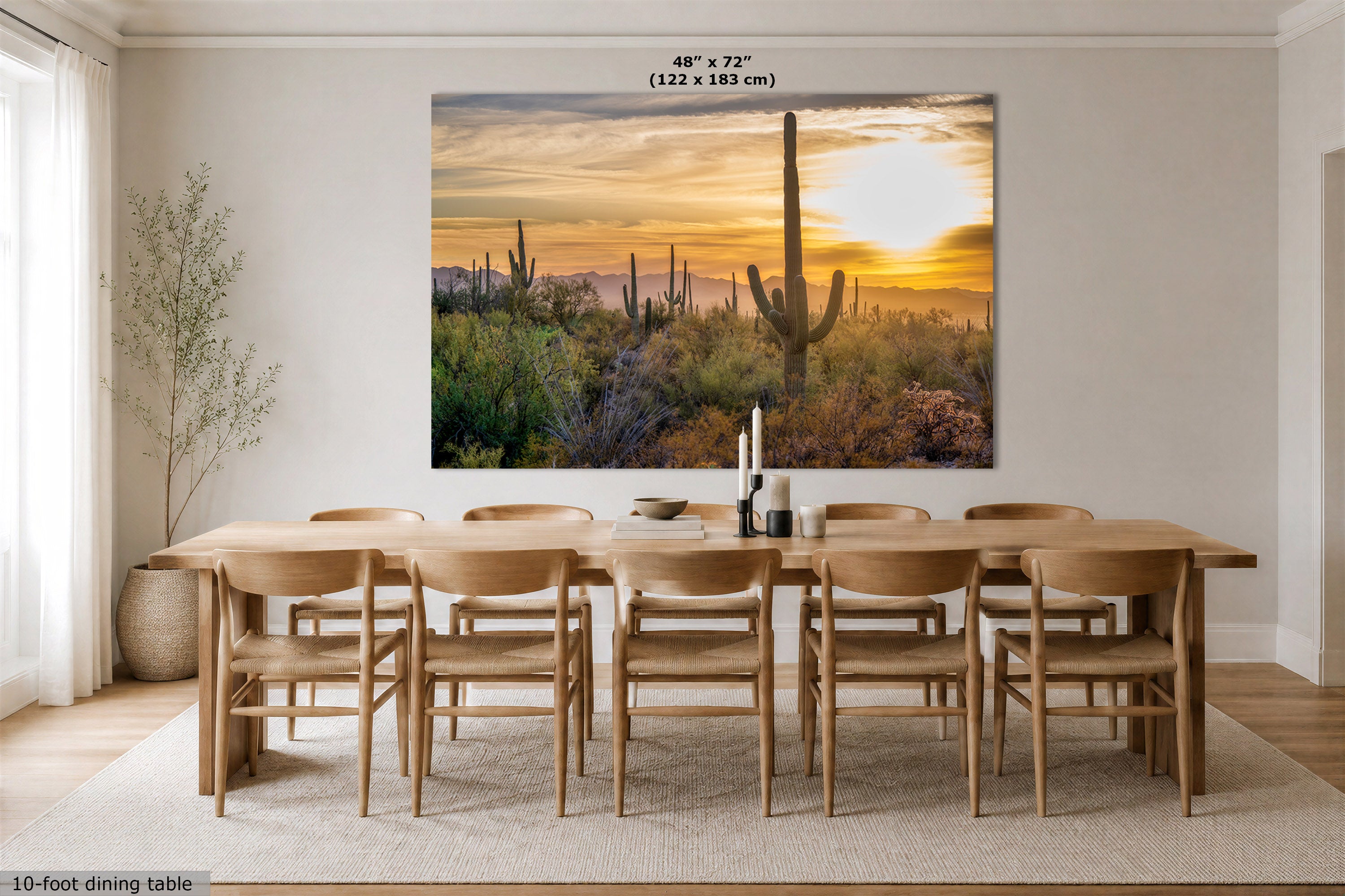 A large rectangular wall art print showing a desert sunset with saguaro cacti, displayed above a wooden dining table and chairs in a bright dining room. The print includes size text at the top and a small note at the bottom left.