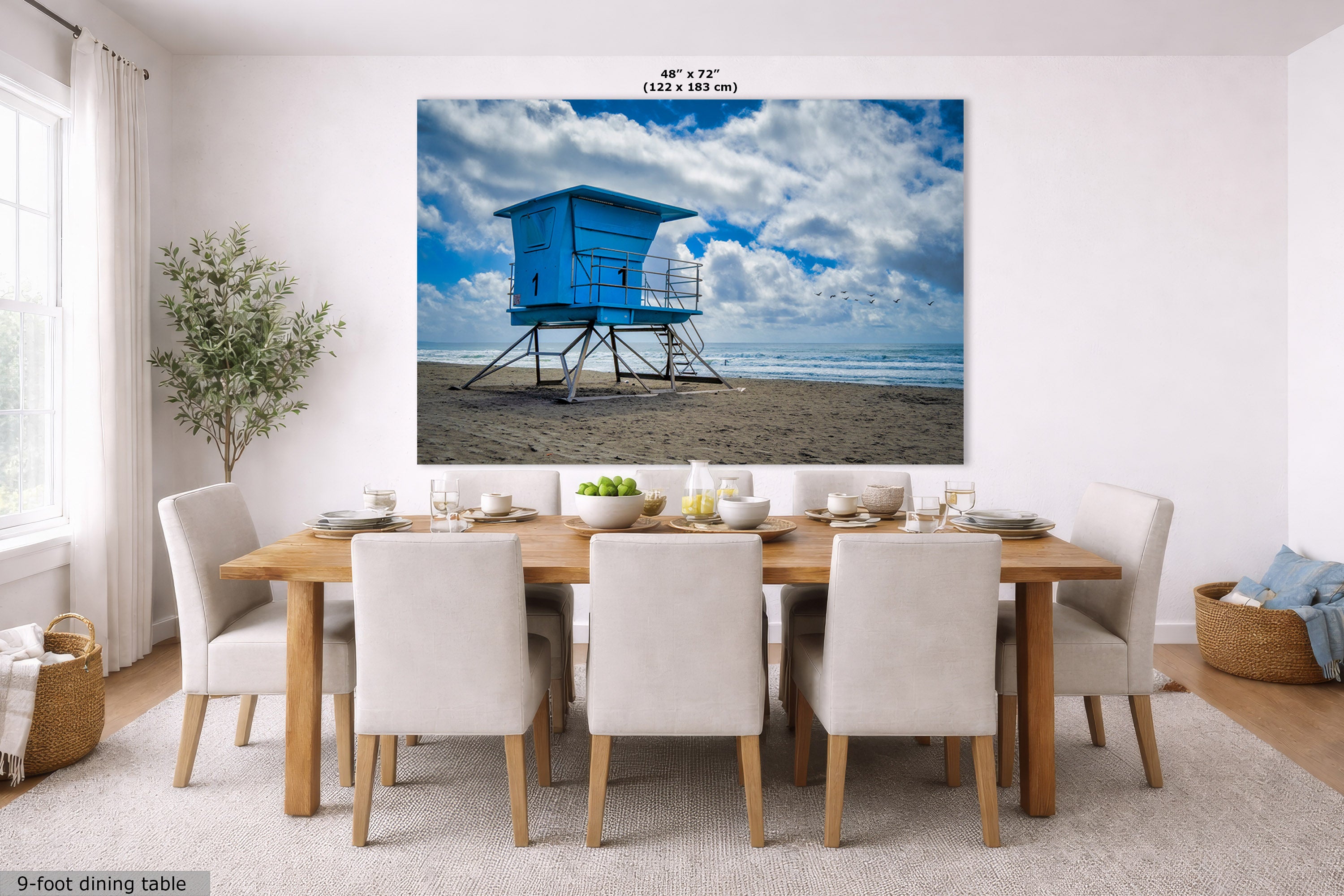 A large rectangular wall art print showing a blue lifeguard tower on a beach under a cloudy sky, displayed above a dining table in a bright modern room. Small text at the top reads 48" x 72".