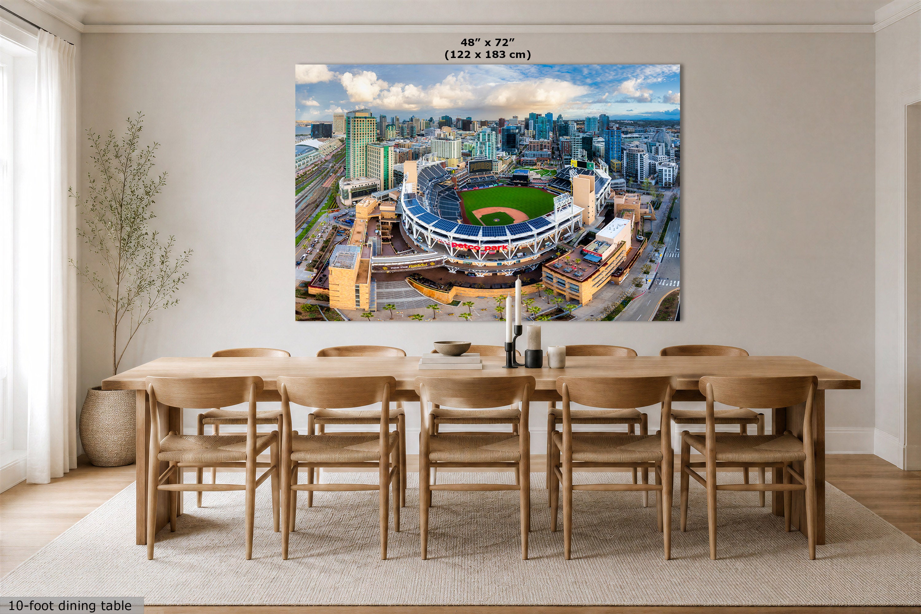 A large rectangular wall art print showing an aerial view of a stadium and city skyline, displayed above a wooden dining table in a bright modern room. Text at the top reads 48 in x 72 in (122 x 183 cm), and small text at the bottom left says 10-foot dining table.