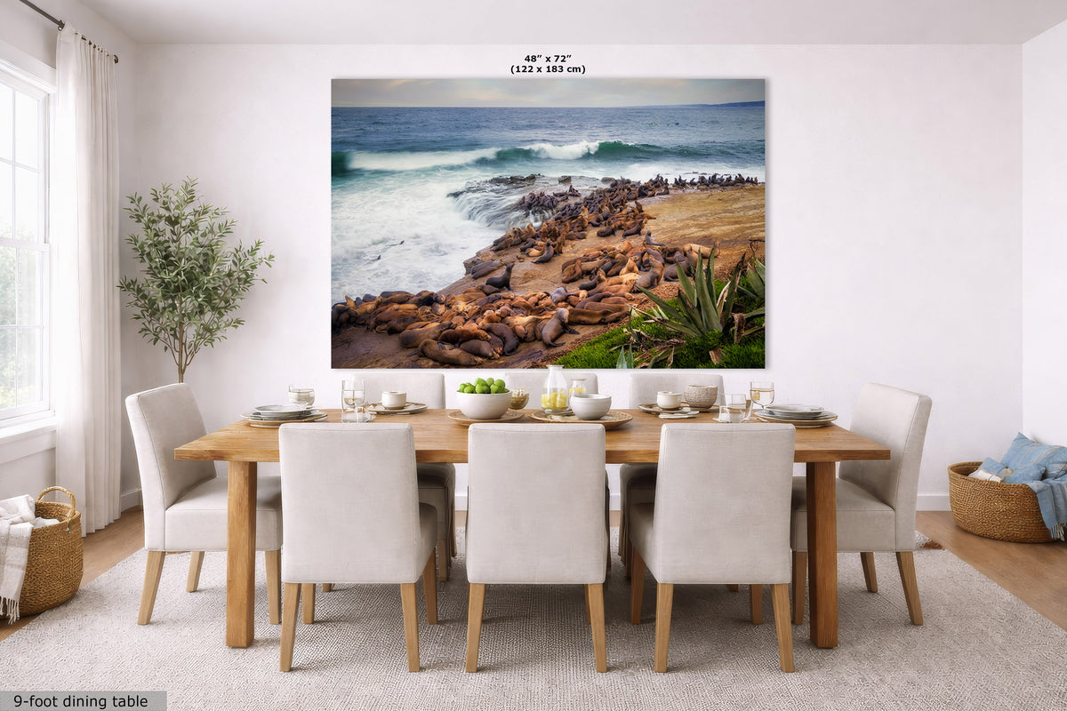 A large wall art print showing a rocky beach and ocean scene, displayed above a dining table in a bright modern dining room. The print includes size text at the top and a small label at bottom left.
