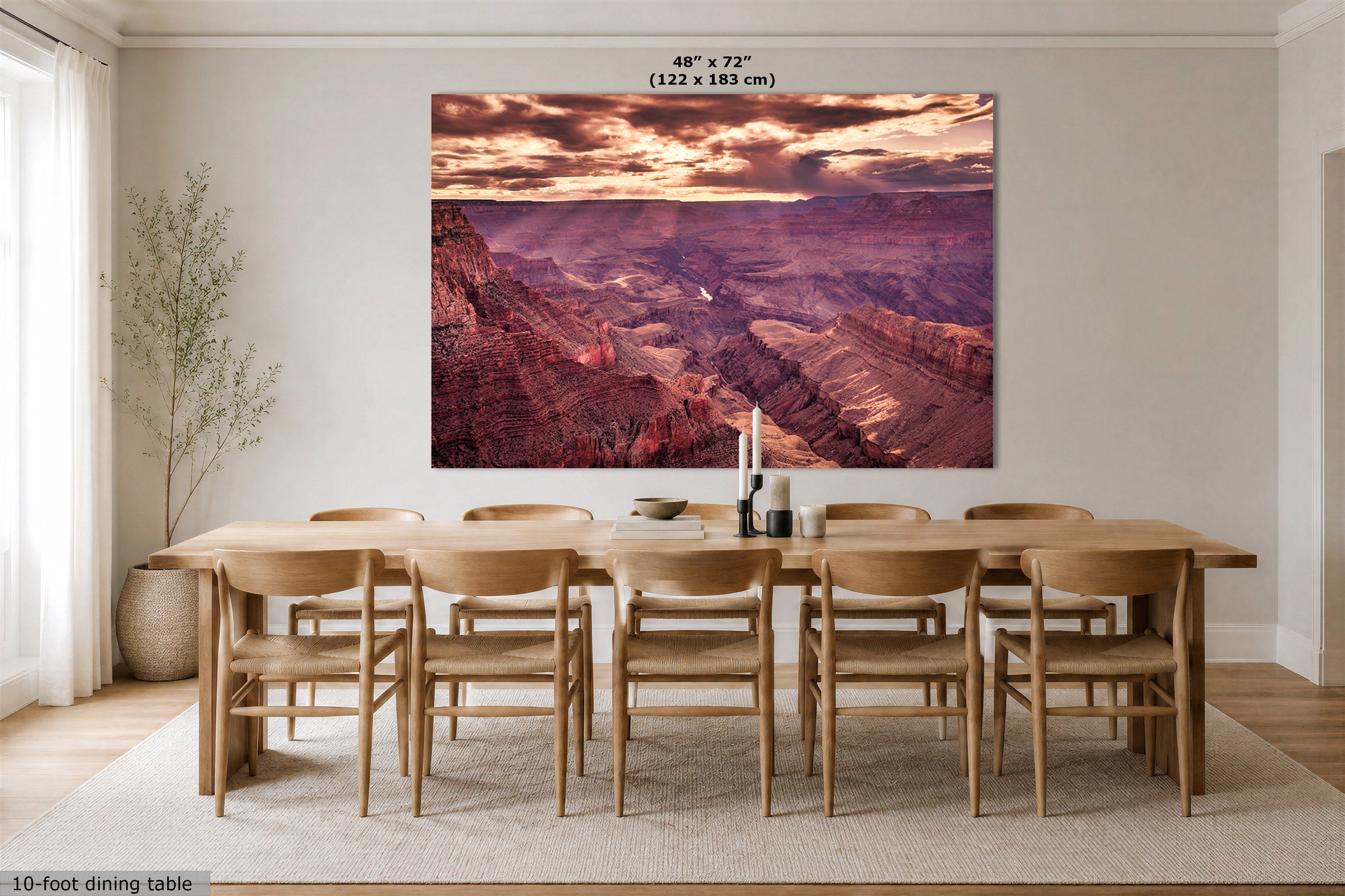 A large rectangular wall art print showing a canyon landscape at sunset hangs above a wooden dining table and chairs in a bright dining room. 