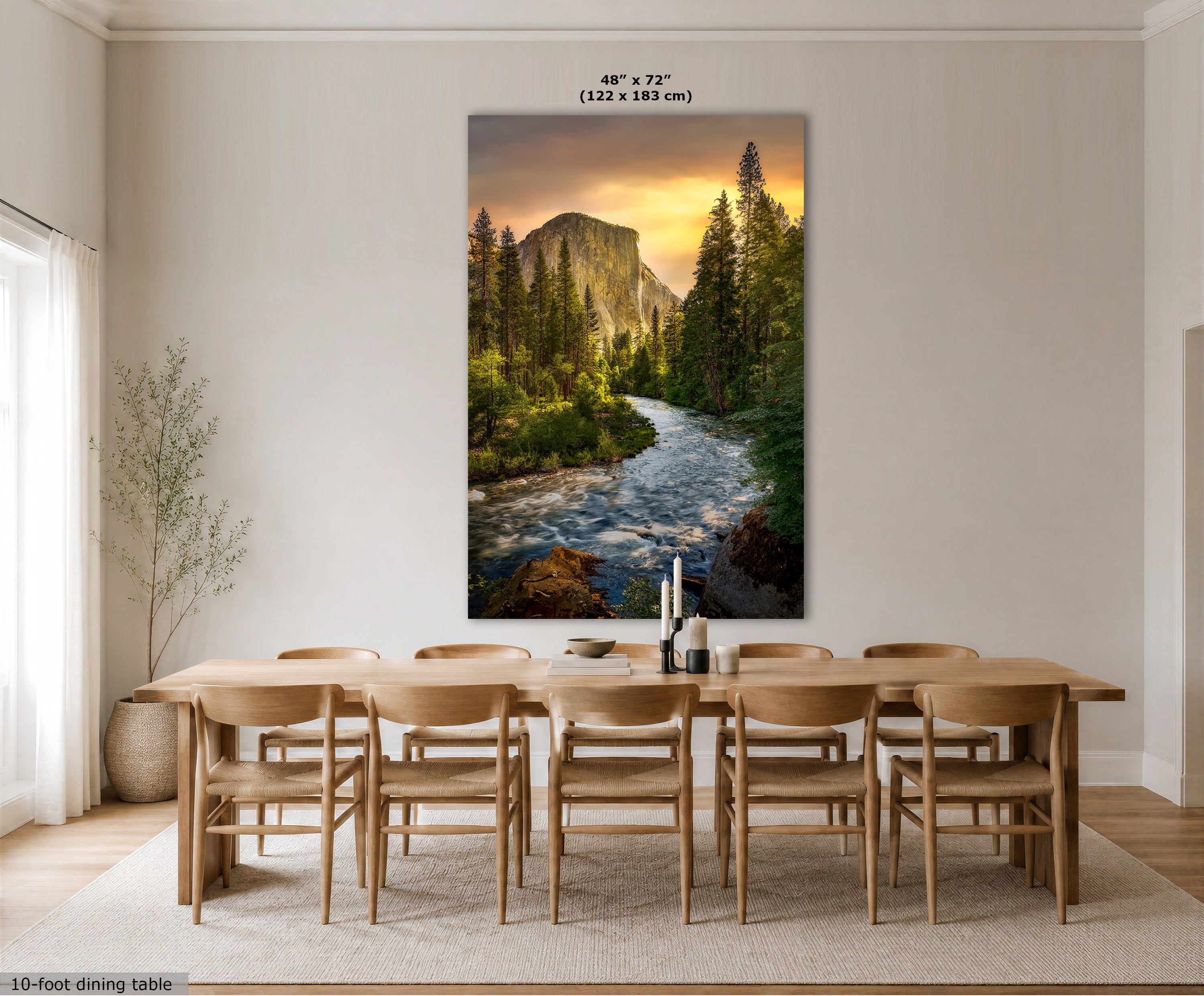 A large vertical El Capitan sunrise wall art print showing a forest stream flowing toward a mountain at sunset, displayed above a wooden dining table and chairs in a bright neutral dining room. The print includes size text at the top and a small size label at the bottom left.