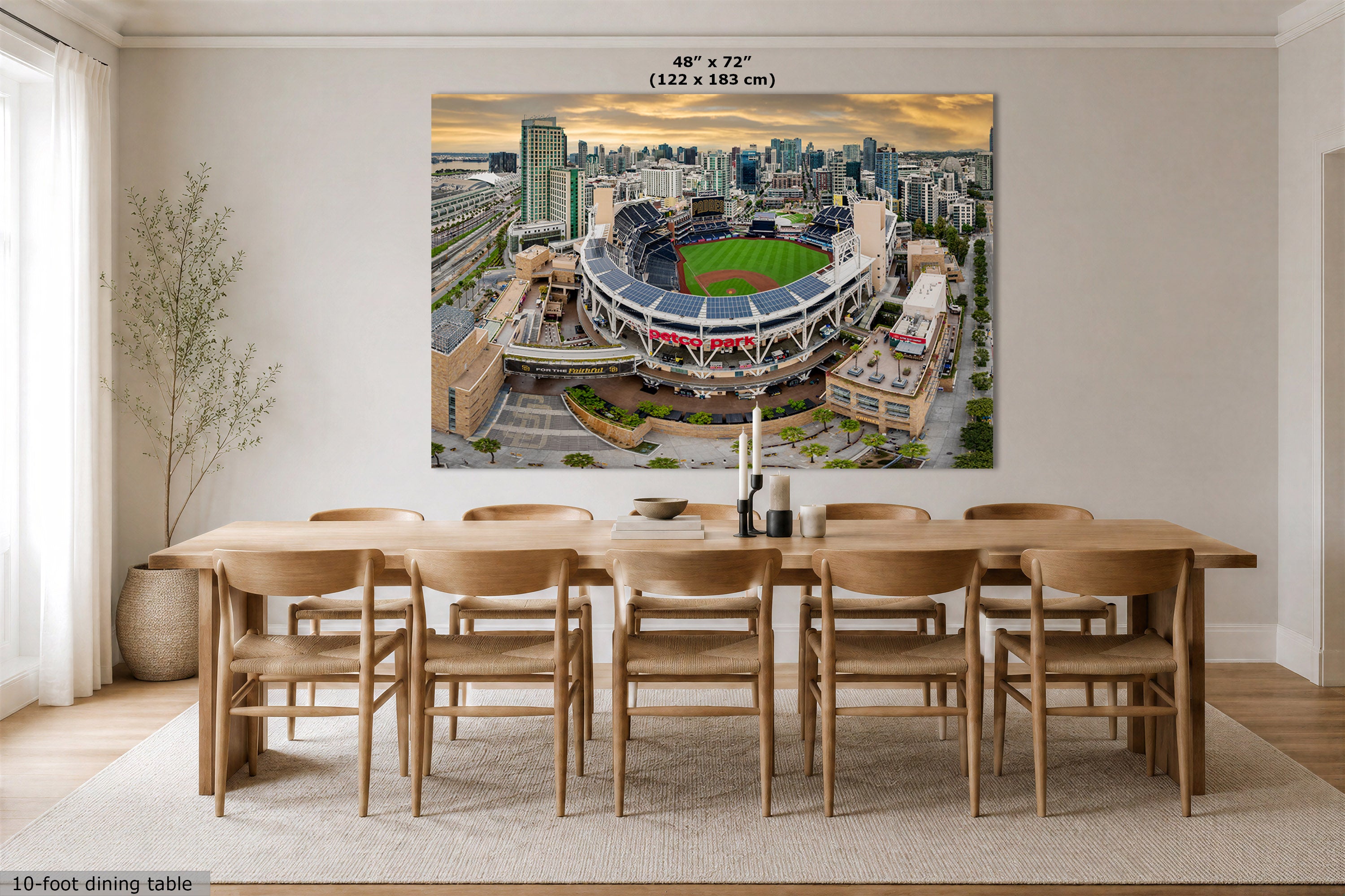 A large wall art print of an aerial stadium scene displayed above a wooden dining table in a modern dining room. The print shows a green field, stadium seating, surrounding buildings, and a sunset sky.