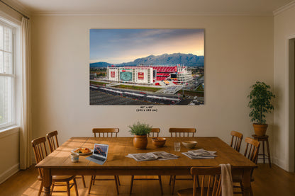 a large, colorful painting of a stadium or arena on the wall of a room with a wooden dining table and chairs.