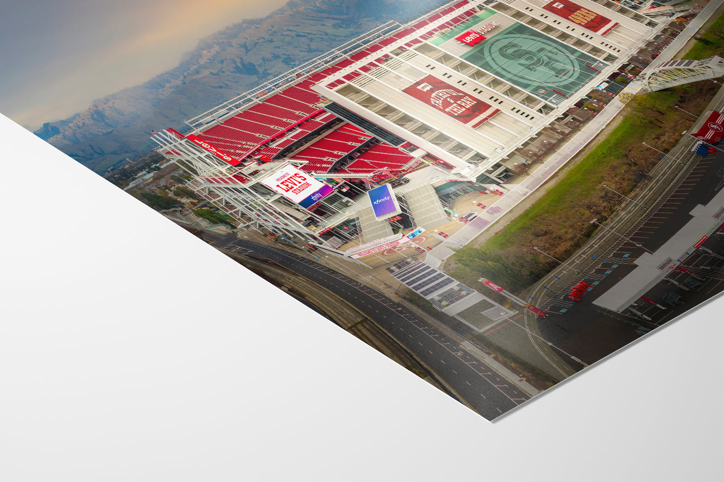The image is an aerial view of a large stadium with a prominent red and white exterior, surrounded by mountains in the background.