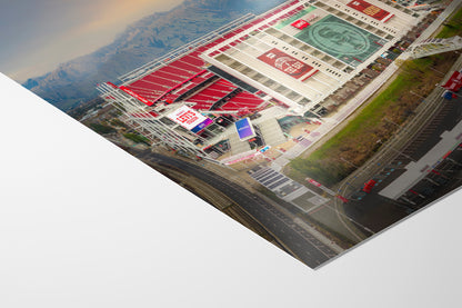 The image is an aerial view of a large stadium with a prominent red and white exterior, surrounded by mountains in the background.