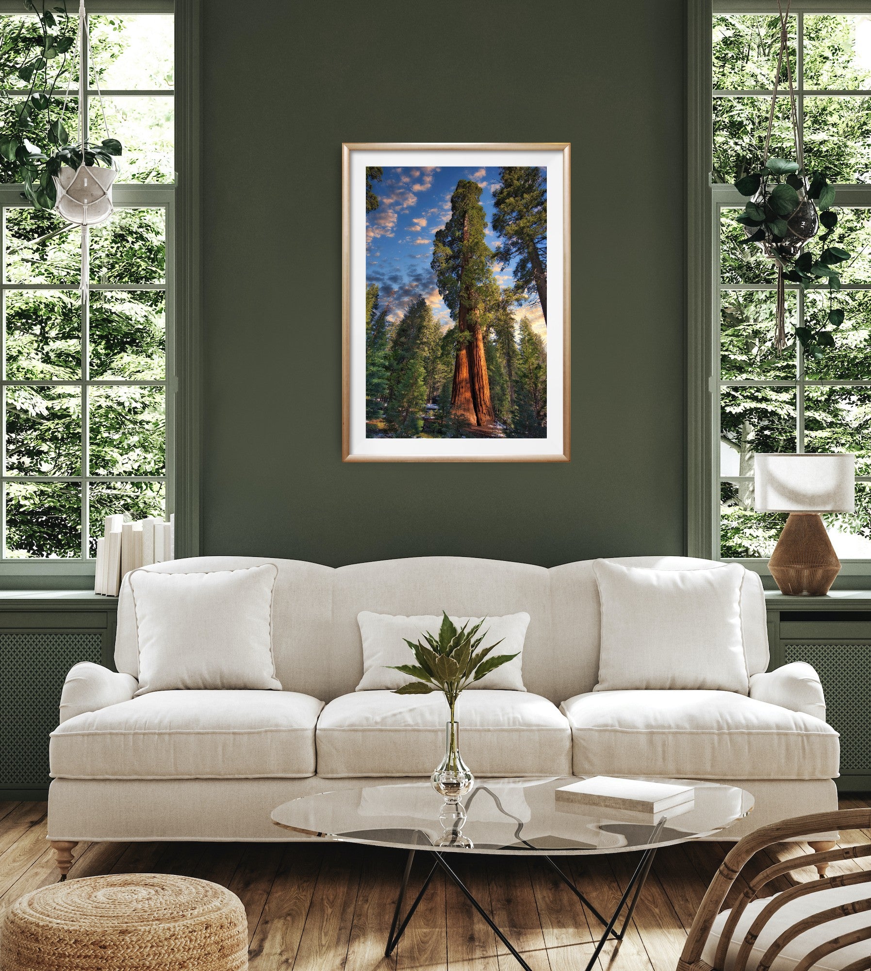 A framed vertical landscape Giant Sequoia art print depicting giant redwood trees under a blue sky, displayed on a dark green wall above a white sofa in a modern living room setting.