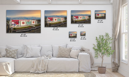 The image is a living room with a white sofa and a potted plant. There are five framed pictures of a stadium displayed on the wall, each with a different size and resolution. The pictures are arranged in a row, showcasing the stadium from various angles and perspectives.