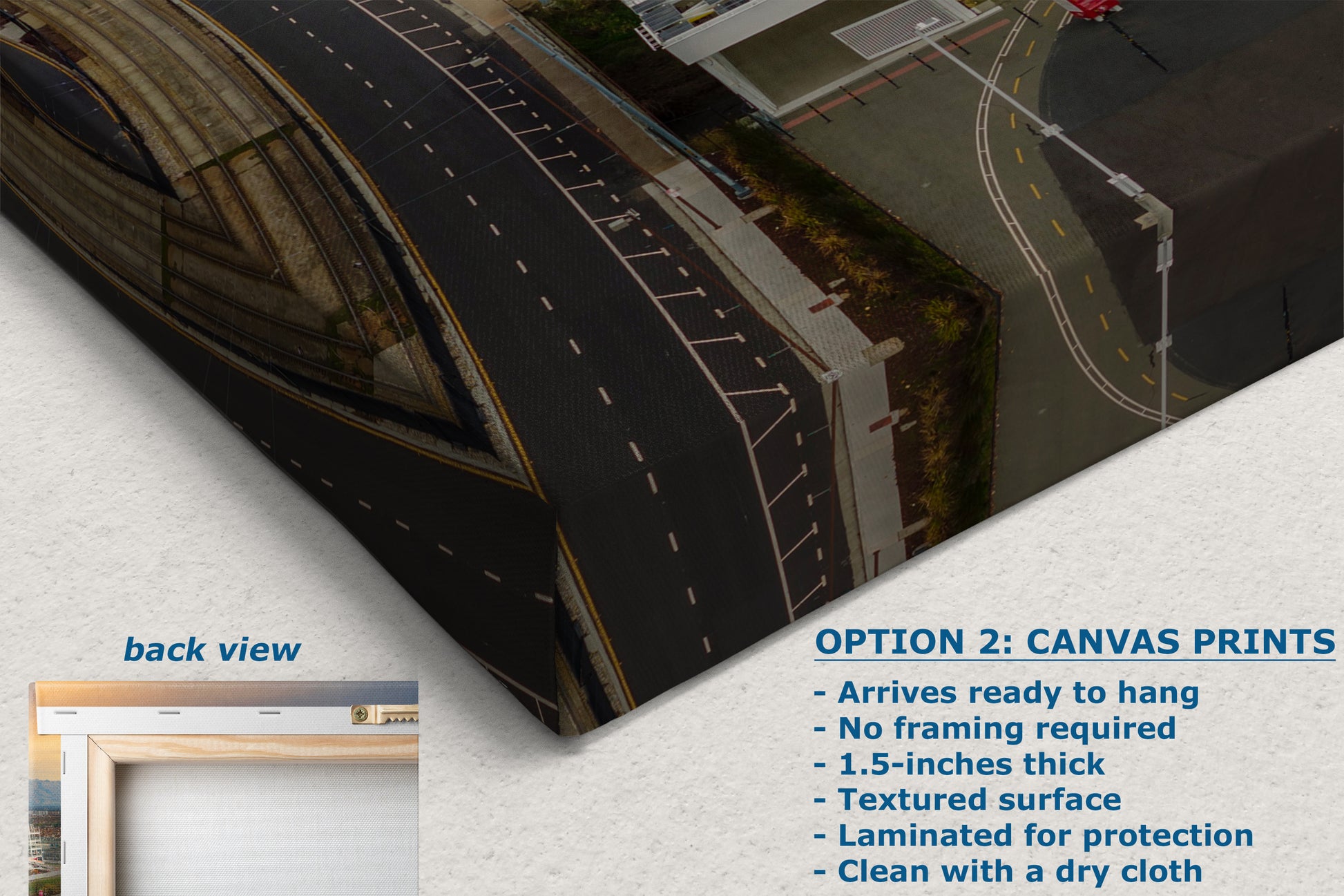 The image is a canvas print featuring an aerial view of a road with buildings and a bridge in the background. The text below the image provides information about the print, including that it is a back view of the print, arrives ready to hang, does not require framing, is 1.5-inches thick, has a textured surface, is laminated for protection, and can be cleaned with a dry cloth.