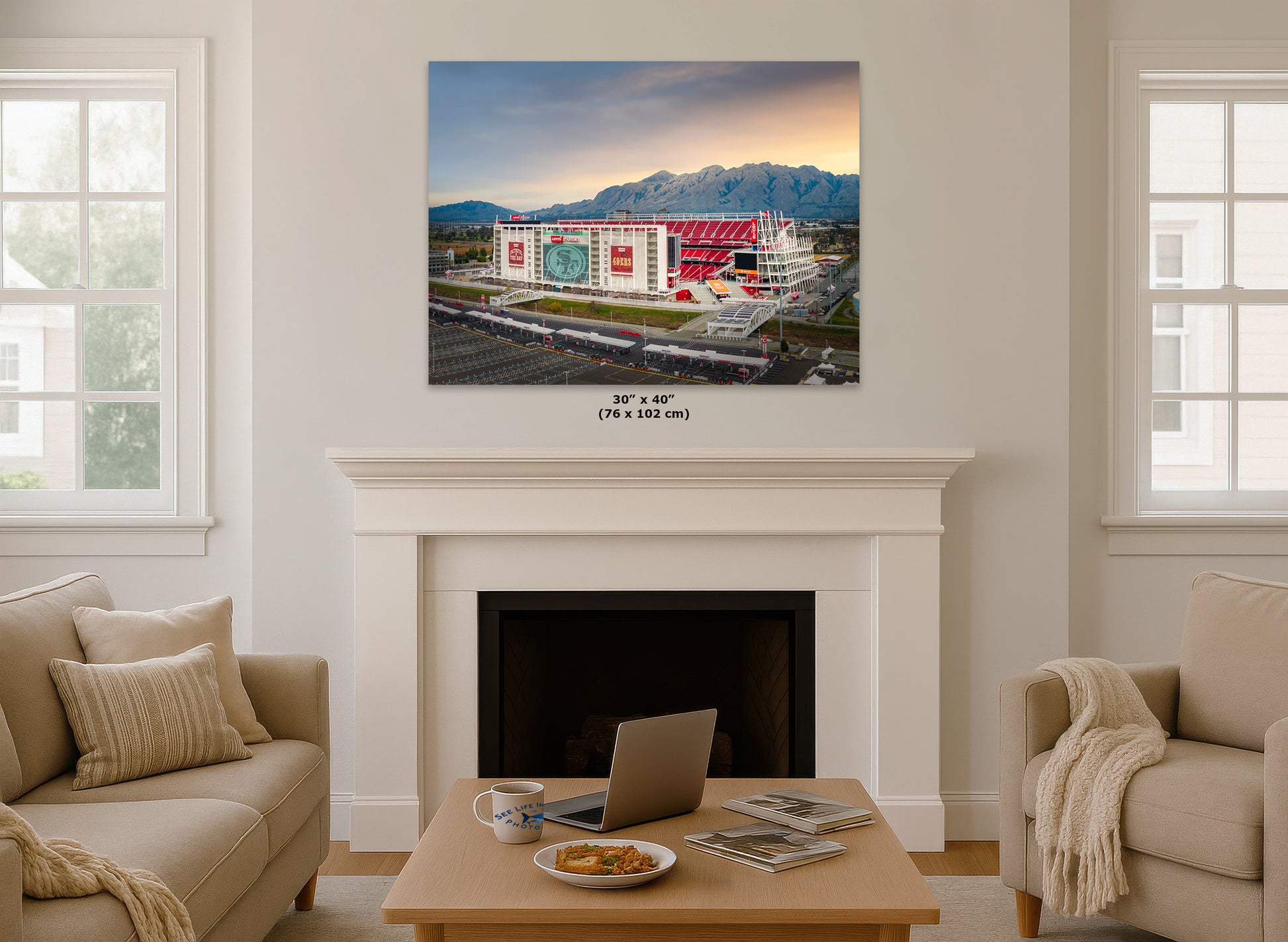 a cozy living room with a fireplace, a coffee table, and two couches. Above the fireplace, there is a large framed picture of a stadium with mountains in the background.