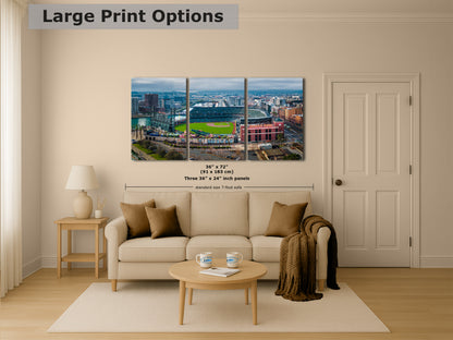 The image is a living room with a large, three-panel print of a cityscape featuring a baseball field in the background. The print is mounted on the wall above a beige sofa and a coffee table.