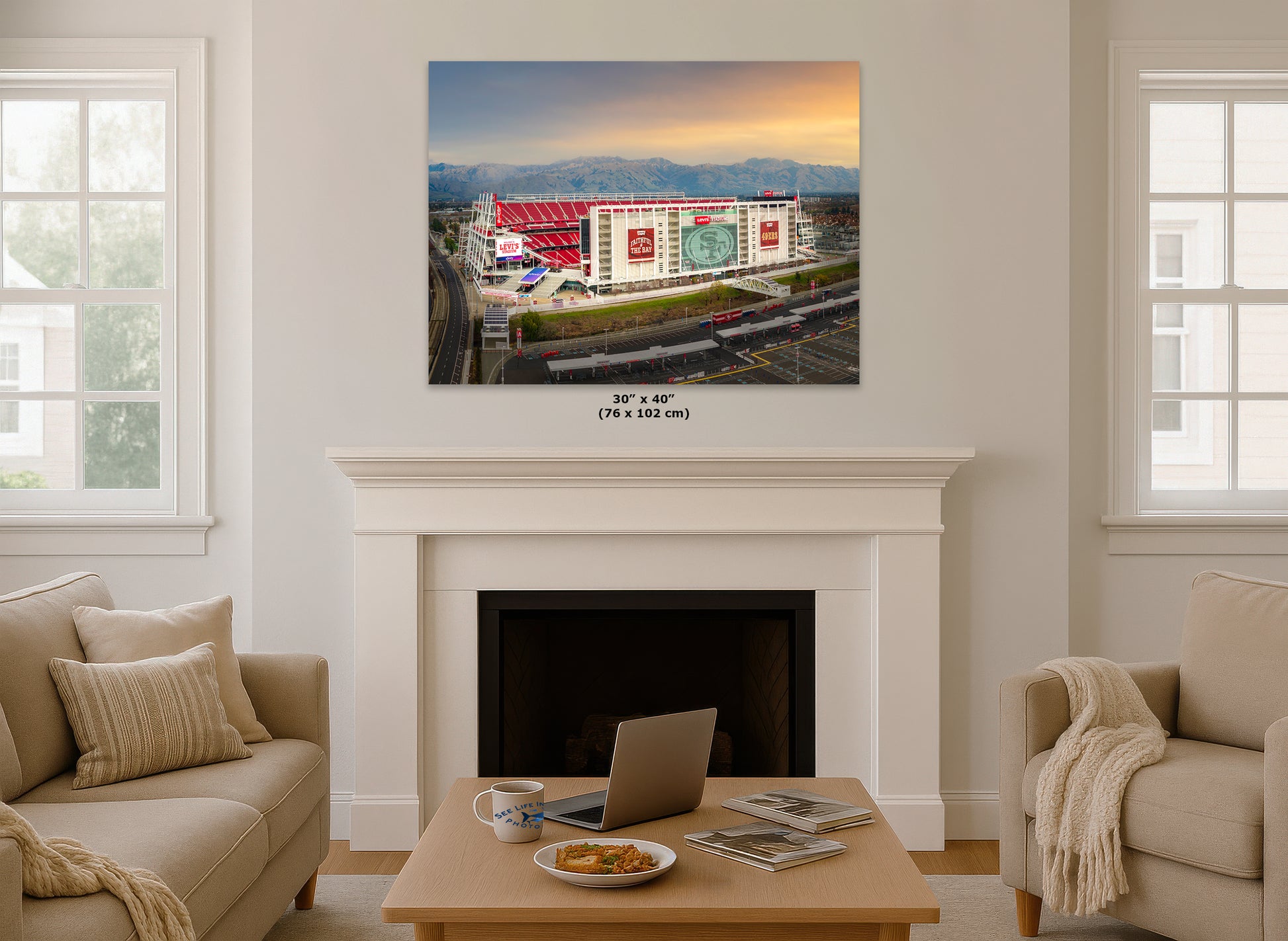 a cozy living room with a fireplace, a coffee table, and two couches. Above the fireplace, there is a framed picture of a stadium with a red roof and white walls.