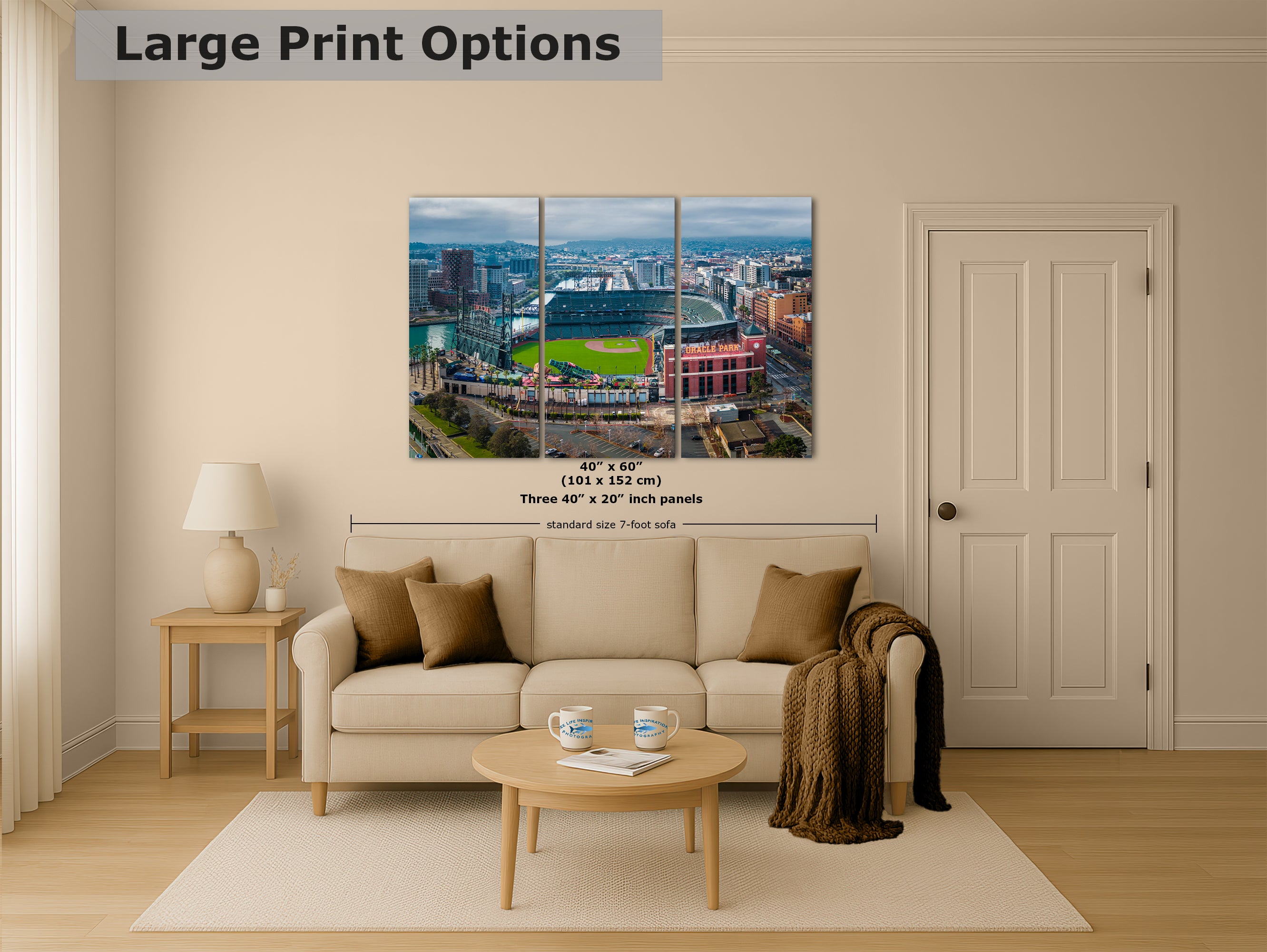 The image is a living room with a large wall art print depicting a city skyline, including a baseball stadium. The print is framed and measures approximately 40 inches by 60 inches, with three panels. The room is furnished with a couch, coffee table, and a lamp, creating a cozy and inviting atmosphere.