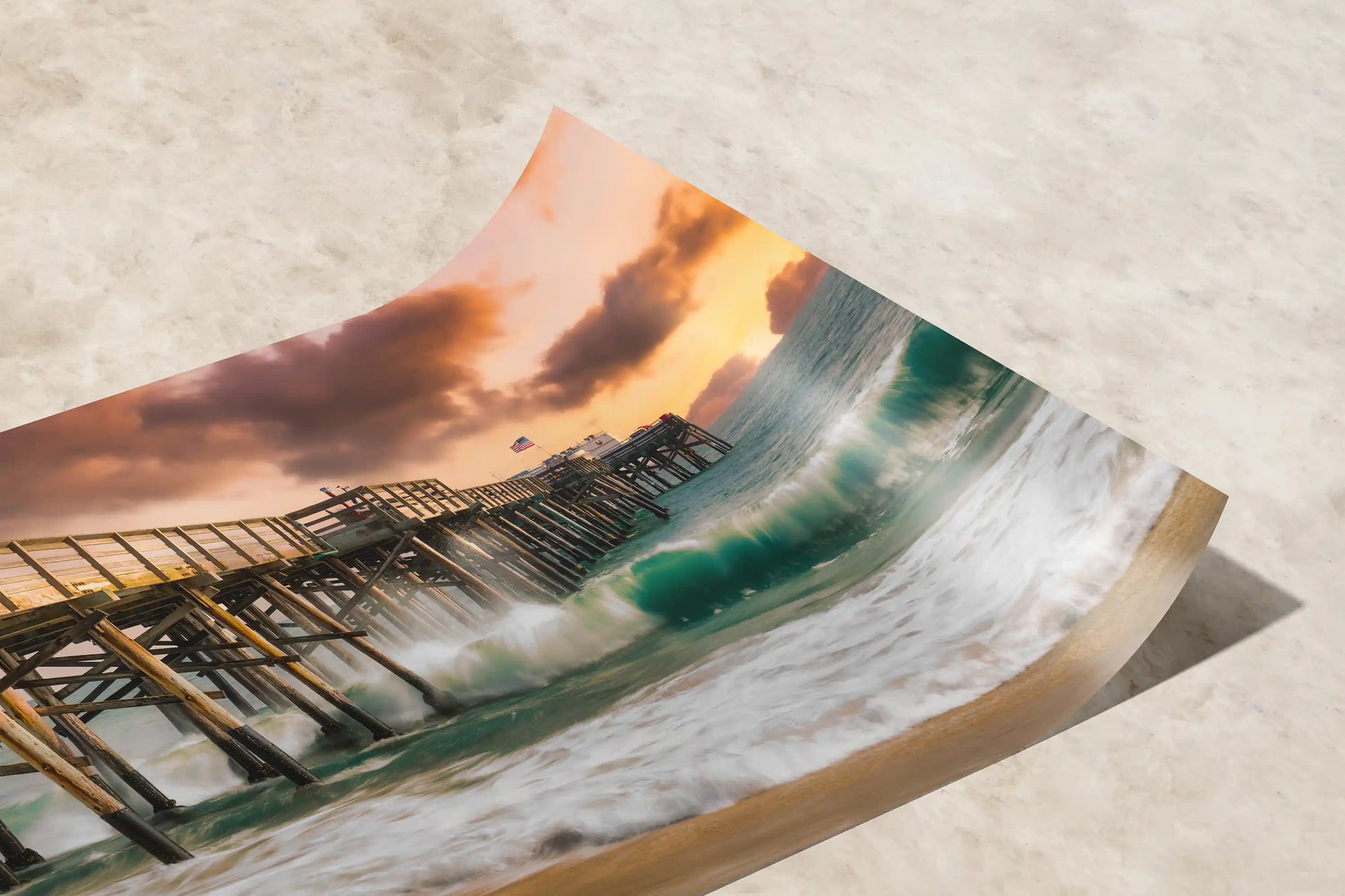 Curled paper print of Balboa Pier in Newport Beach with a vibrant wave cresting under a fiery sunset sky, perfect for coastal-themed decor.