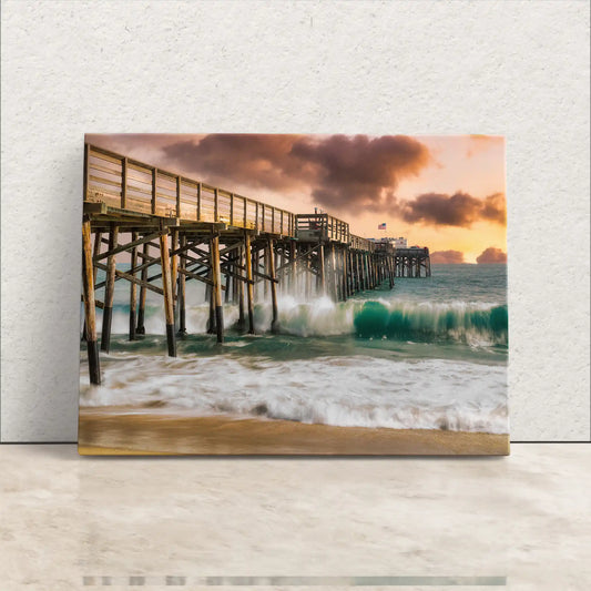A gallery-wrapped canvas print of Balboa Pier at sunset, with waves crashing against the pillars and a vibrant sky.