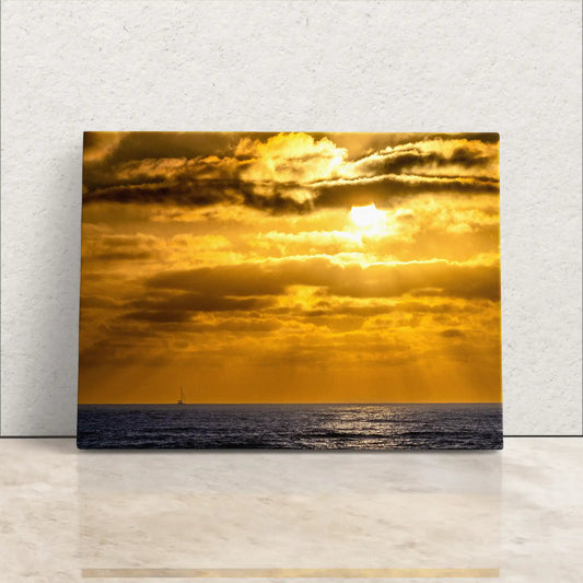 A canvas print leaning against a white wall, featuring a vibrant California sunset with golden hues and a distant sailboat on the horizon.