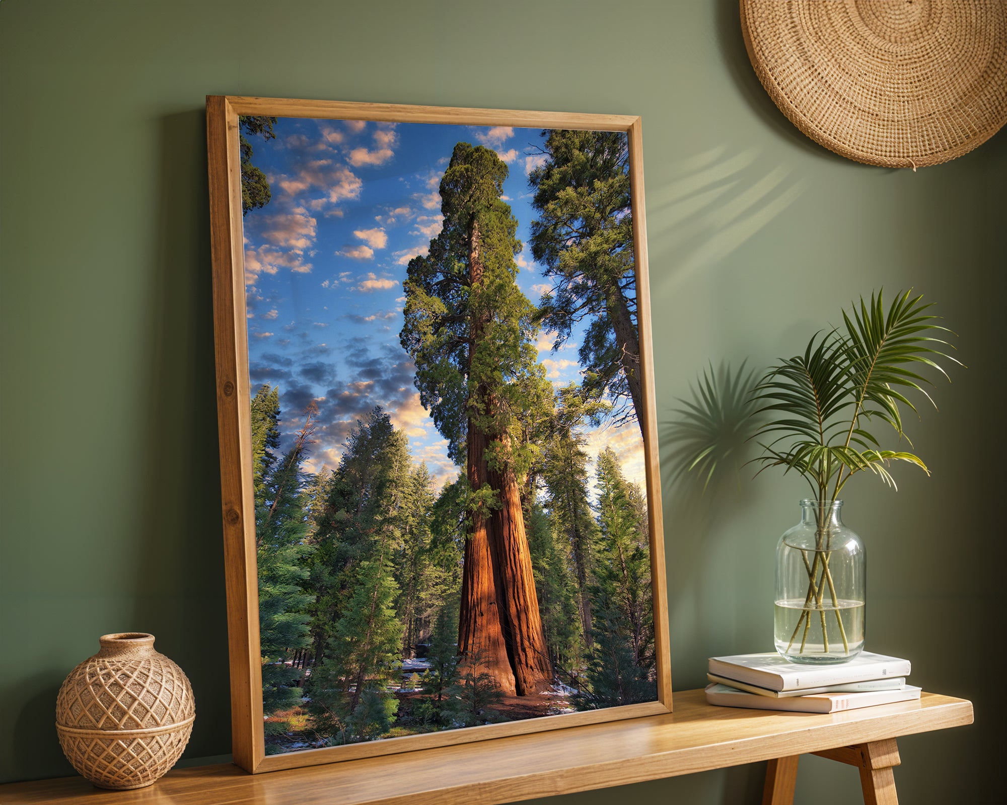 A framed vertical Giant Sequoia art print depicting a sequoia tree in a forest under a blue sky, styled on a wooden console table in a green room with a woven vase and a plant.