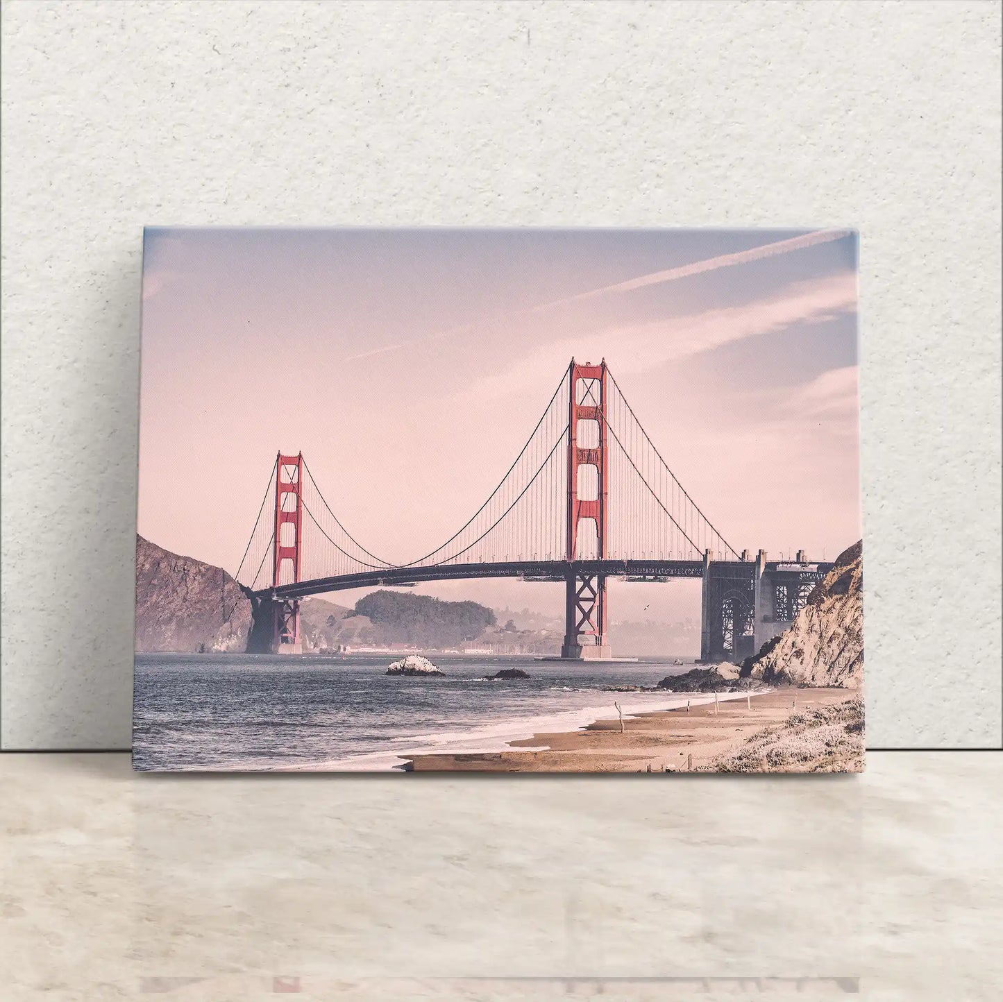 A canvas print leaning against a wall with a vintage-style photograph of the Golden Gate Bridge in soft pink hues, with a clear sky and calm waters.