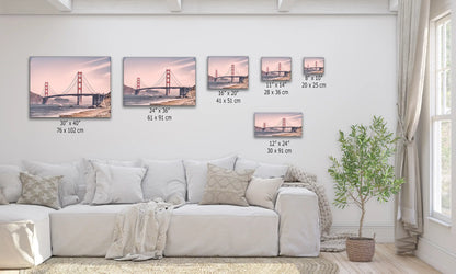 Multiple sized canvas prints of the Golden Gate Bridge, arranged above a sofa to show scale, with the bridge bathed in pink tones at sunset.
