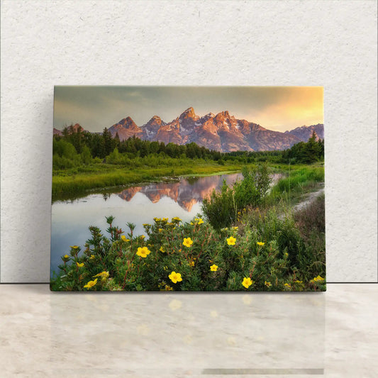 Front view of a canvas print leaning against a white wall, depicting the Grand Teton Mountains at sunrise with foreground wildflowers.